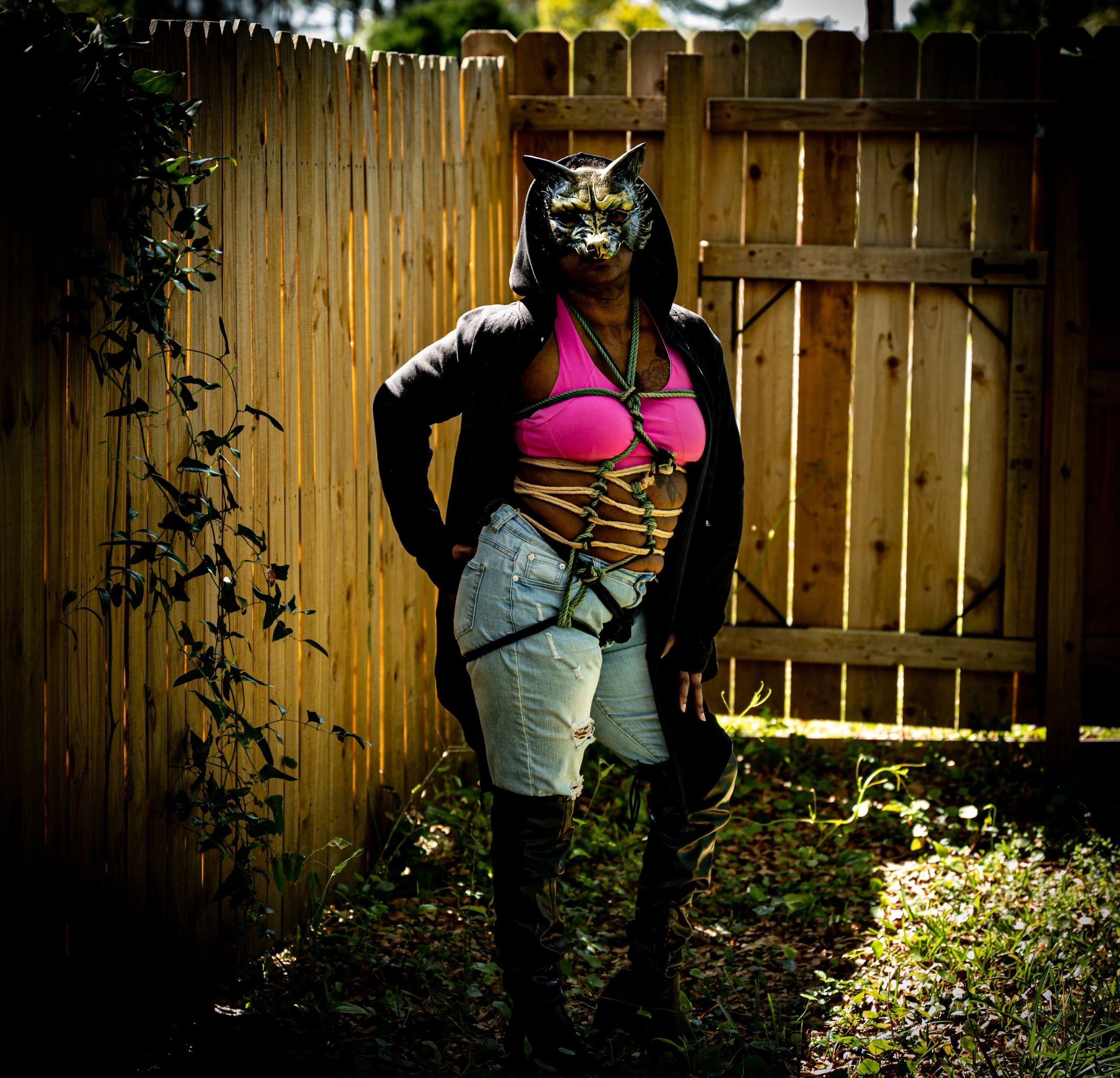 Person standing in a backyard wearing a black cat mask, pink bra, black hoodie, ripped jeans, and knee-high black boots. They are tied with rope in a bondage style around their body.
