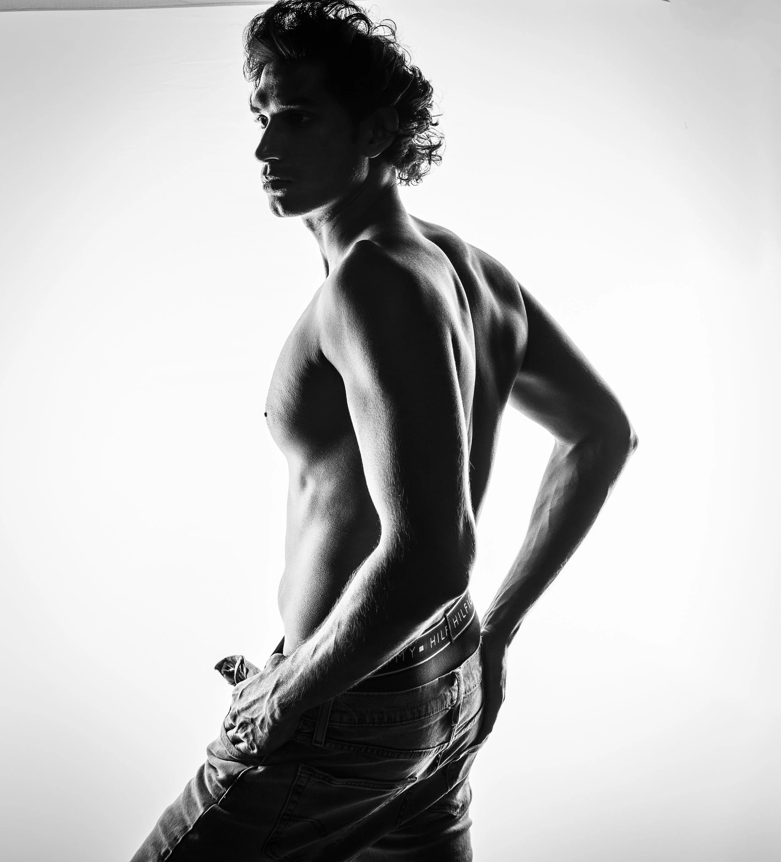 Black and white photo of a shirtless man with curly hair, wearing jeans and Tommy Hilfiger underwear, standing against a plain light background in a studio setting.