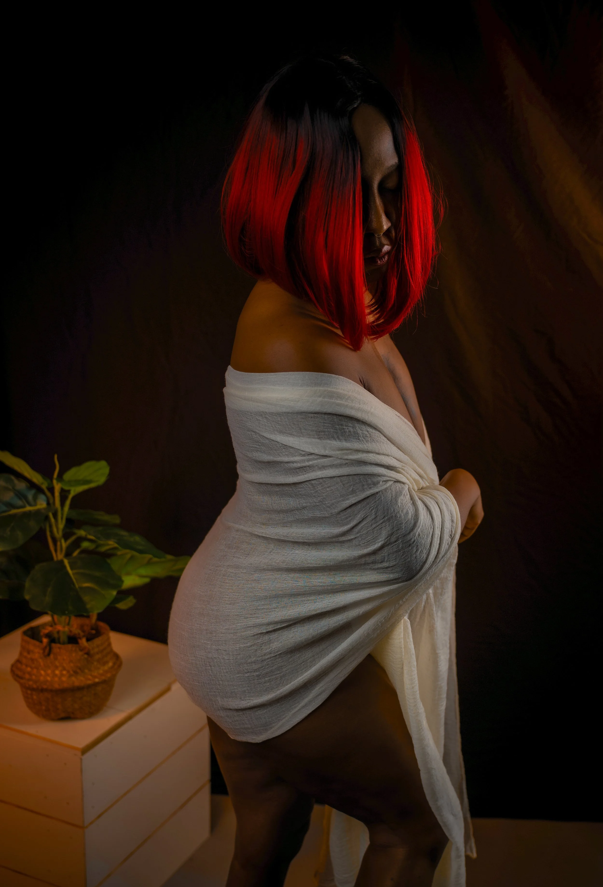Woman with black and red hair wrapping a white towel around her body, standing against a dark background with a potted plant on a wooden table nearby.