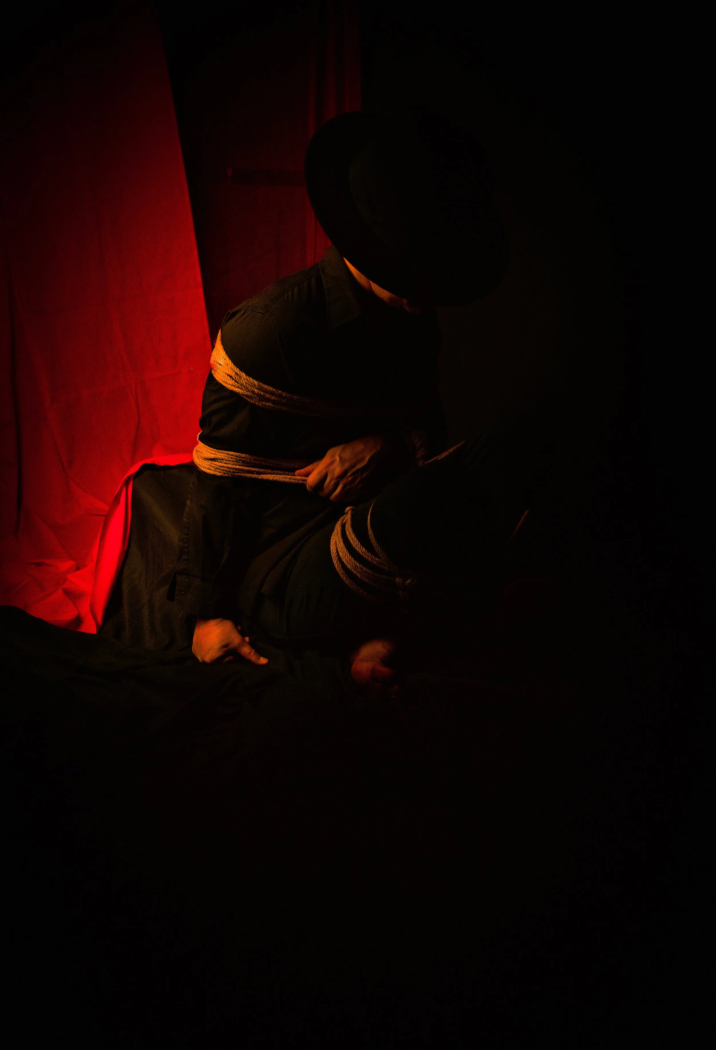 A person in dark clothing and a wide-brimmed hat sits with their torso hunched forward, arms crossed, against a red curtain background. The scene is dimly lit.