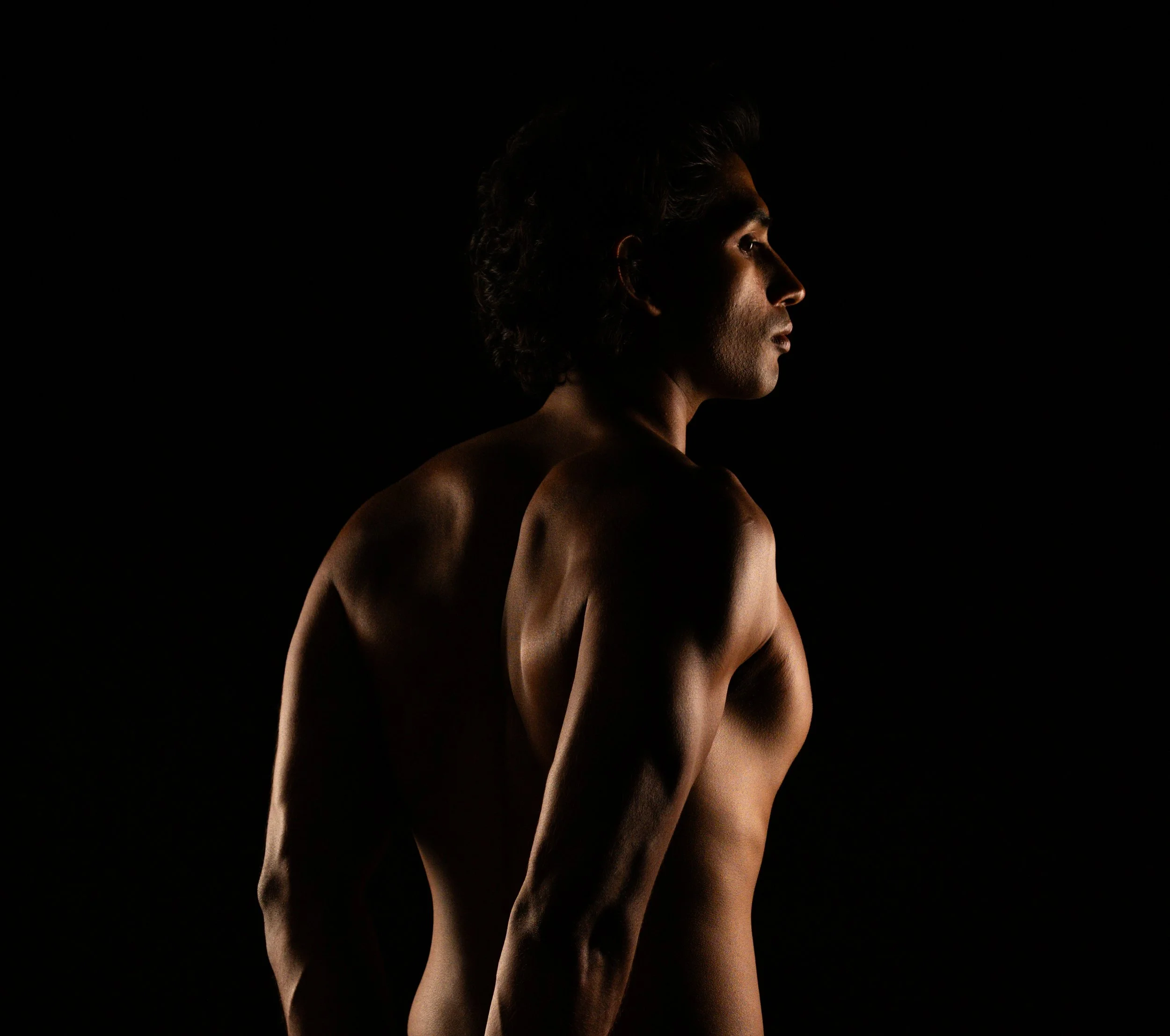Dark studio portrait of a shirtless muscular man shot in profile, with warm directional lighting sculpting the back and shoulders against a black background.