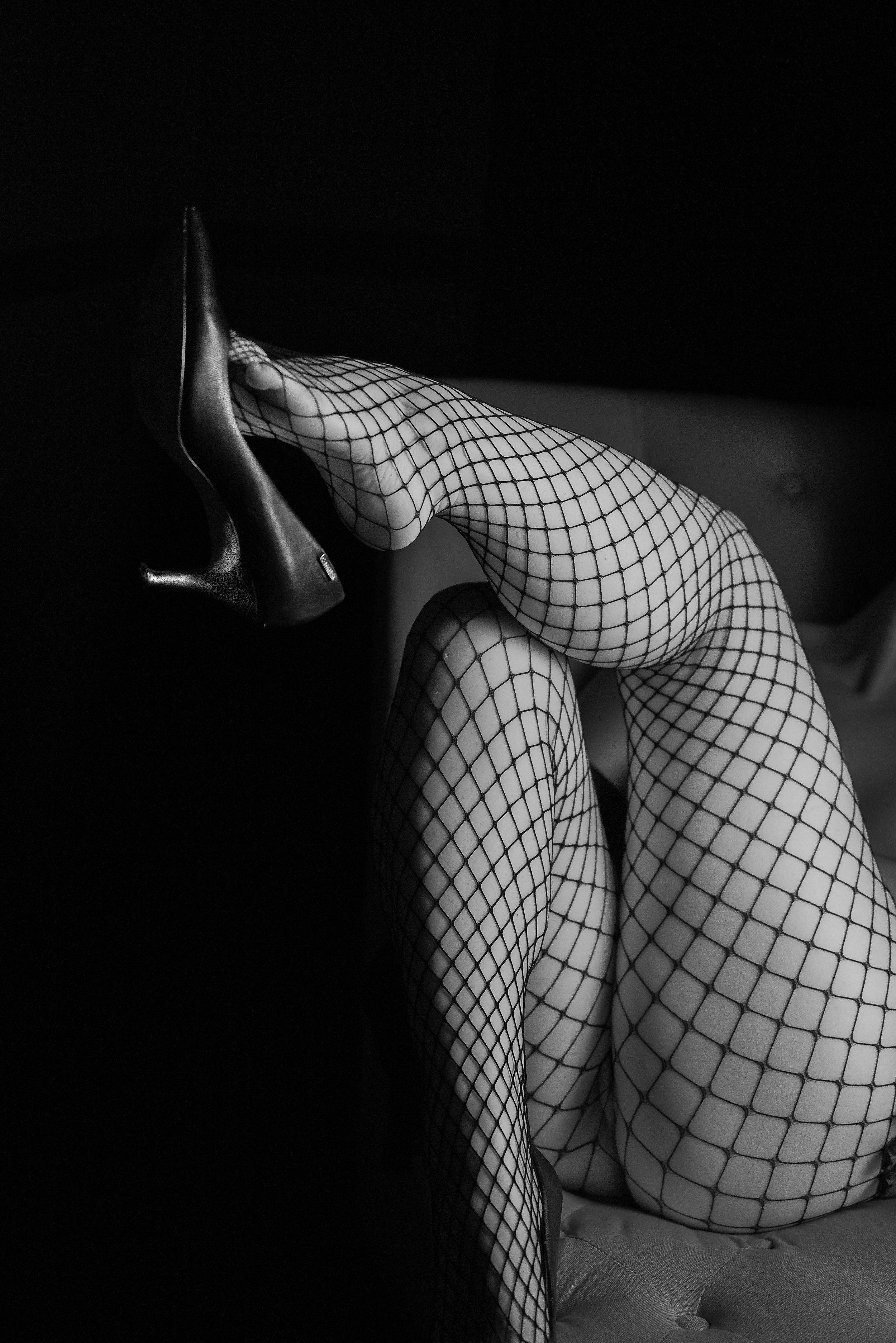 Black and white photograph of a person wearing fishnet stockings, with one leg resting on a surface and the other slightly bent, in a dark setting.
