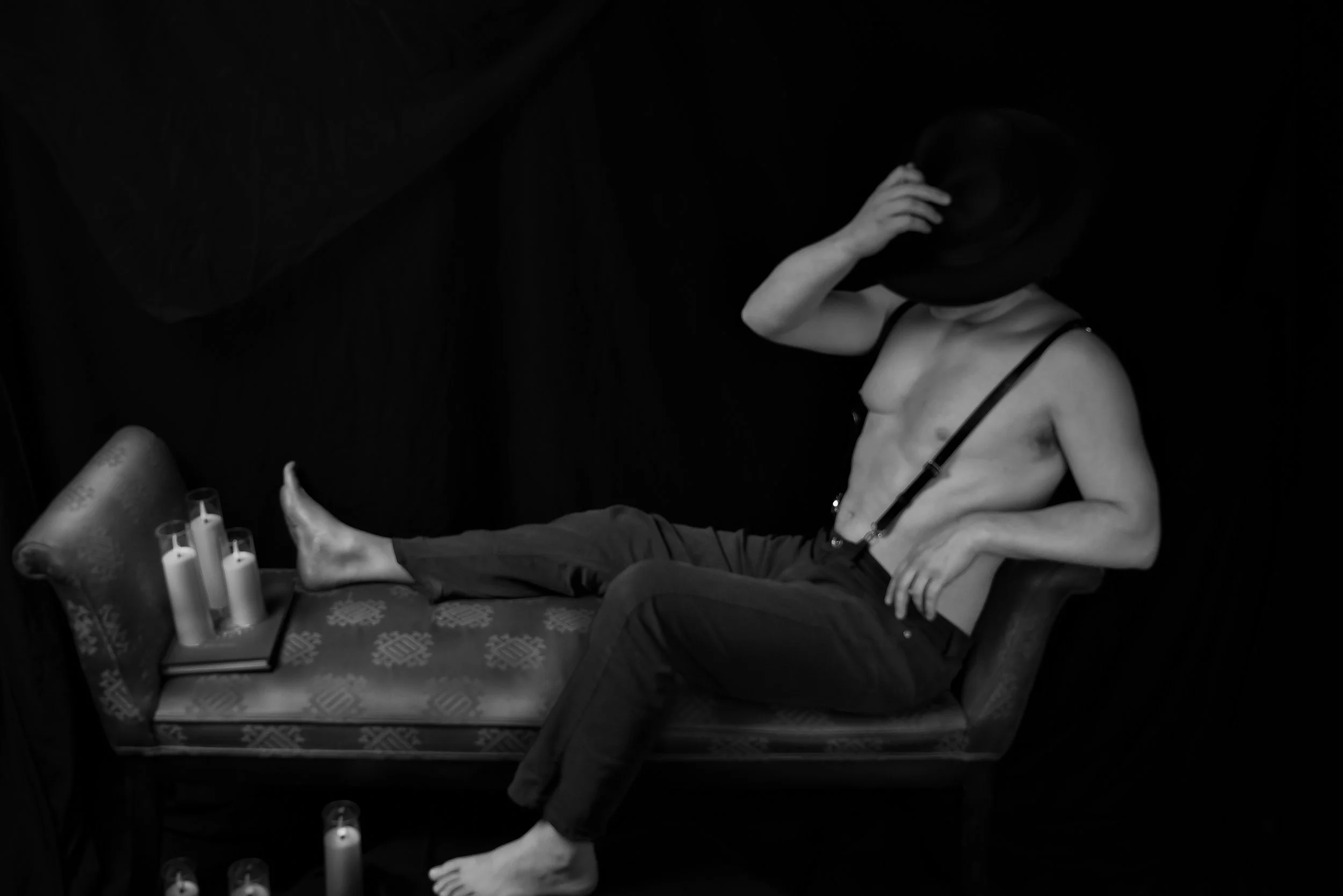 A shirtless man with suspenders sitting on a vintage bench with candles nearby, holding a hat to his face against a dark background.