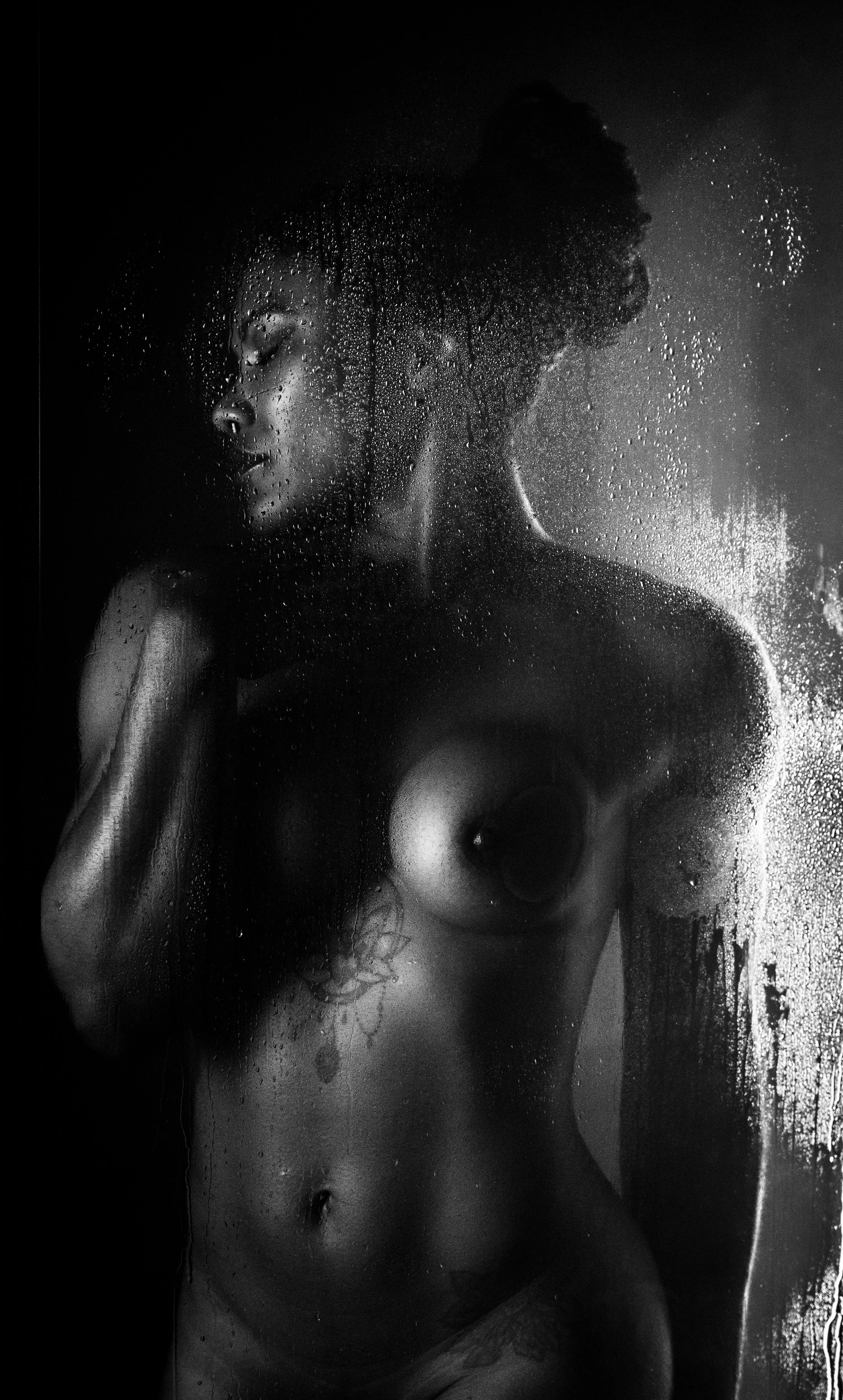 Dramatic black and white fine art nude. Glistening skin catches sharp side light against dark, rain-streaked glass. High contrast shadows sculpt a powerful, timeless silhouette.