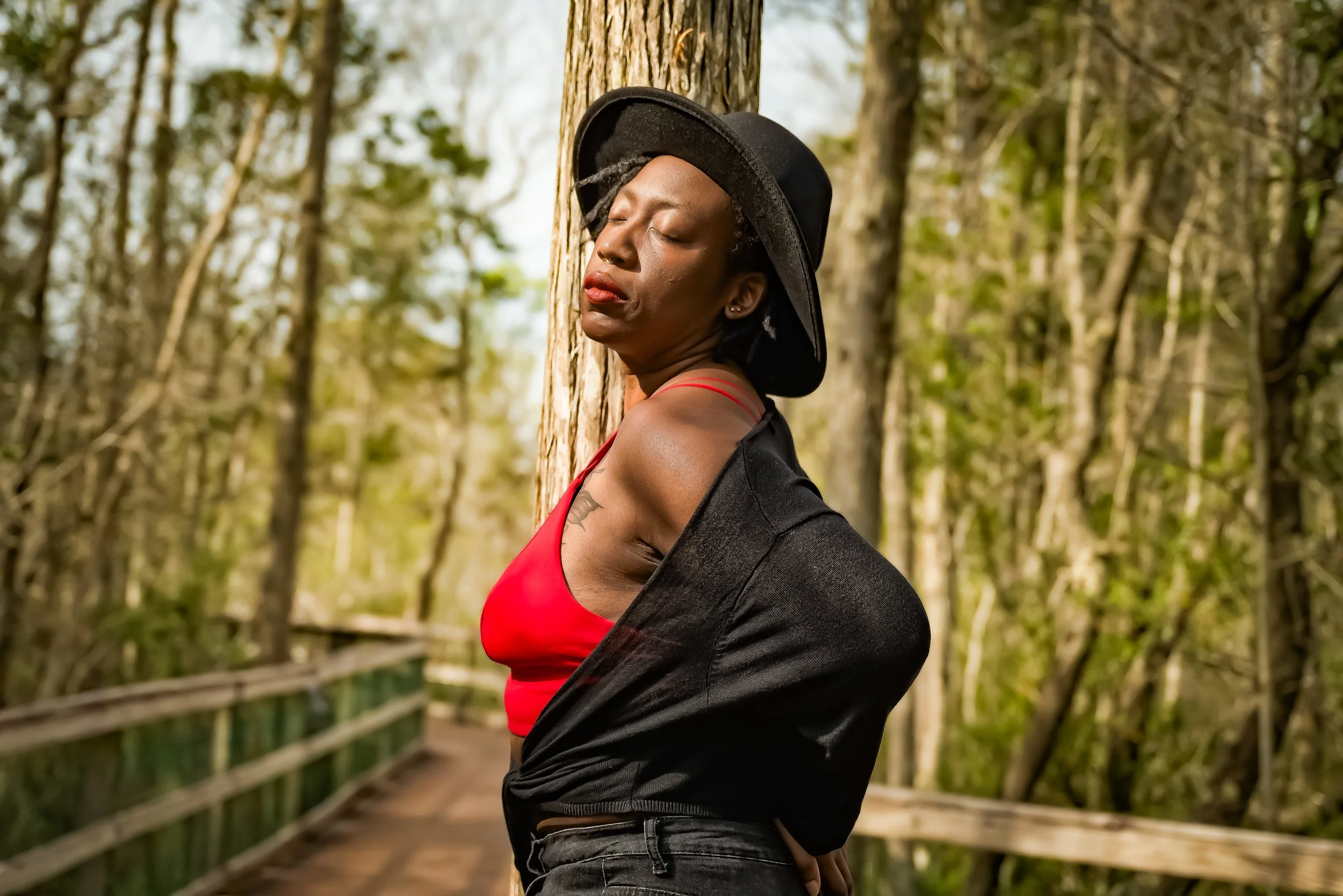 A woman with dark skin and short curly hair wearing a black hat, red top, and black jacket, standing with eyes closed against trees on a bridge in a forested area.