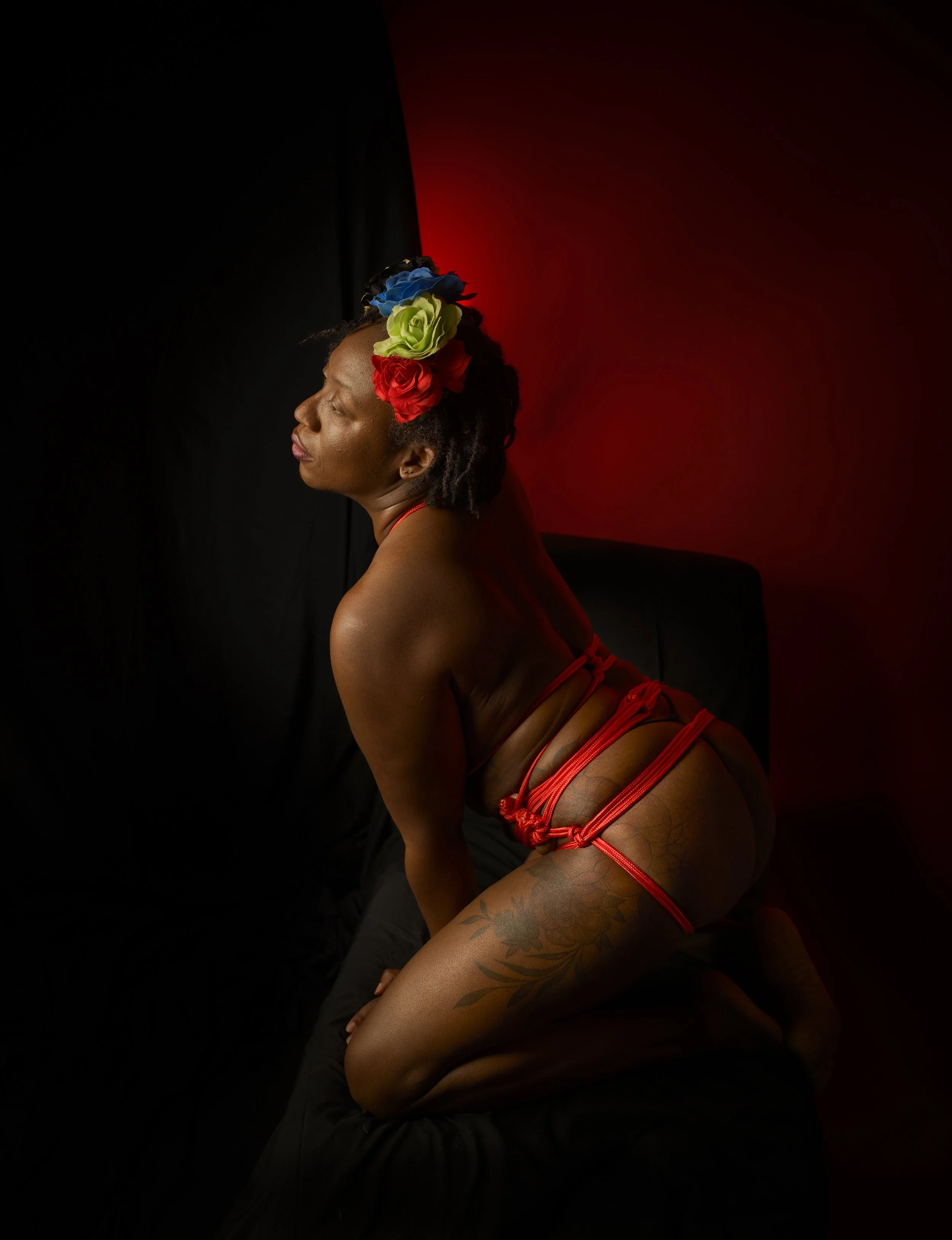 A woman with flowers in her hair, wearing red string lingerie, kneeling on black fabric with a dark background and red lighting to the right.