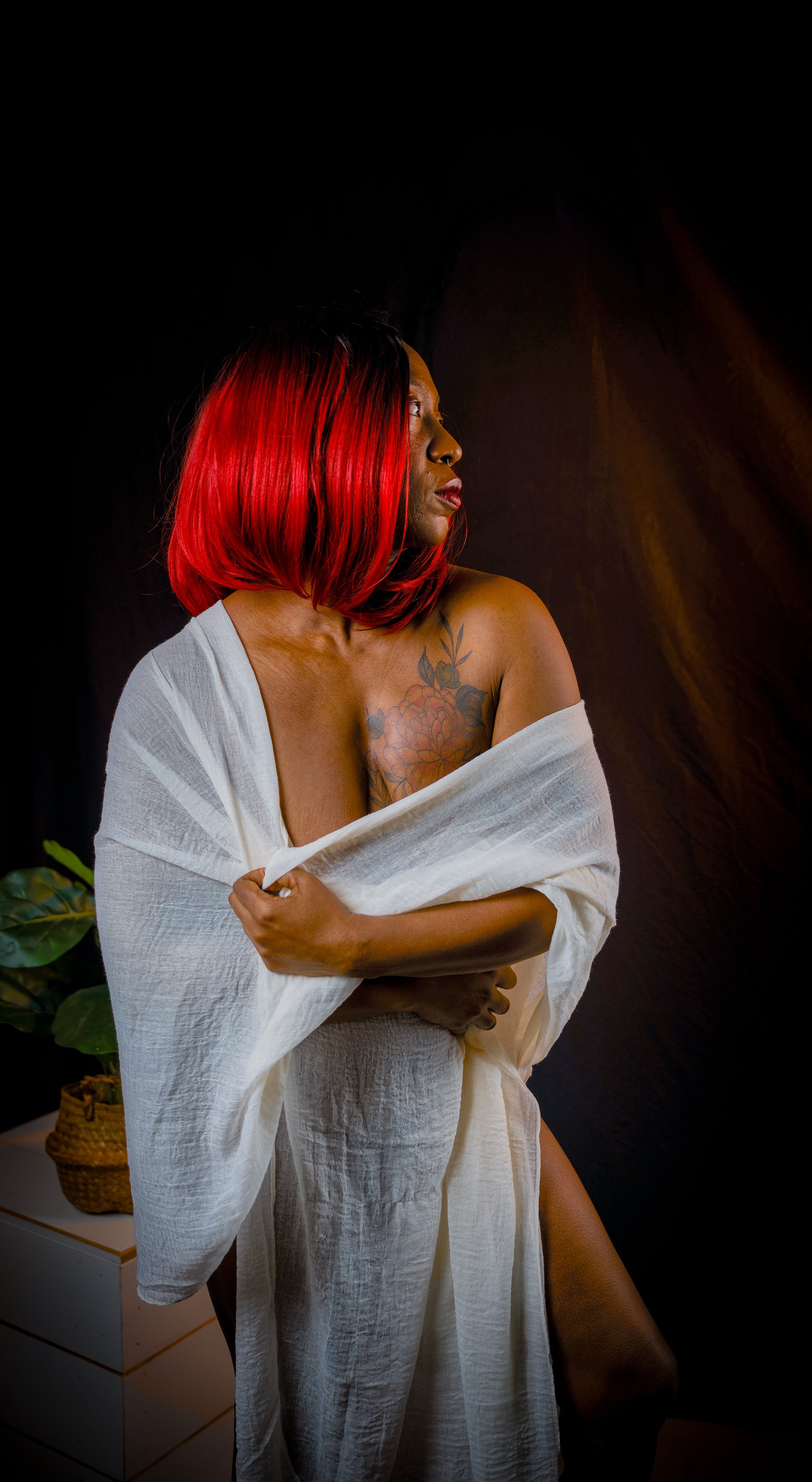 A woman with shoulder-length red and black hair, partially wrapped in a white towel, looking to her right with a tattoo of flowers on her chest, standing next to a small nightstand with a potted plant against a dark background.