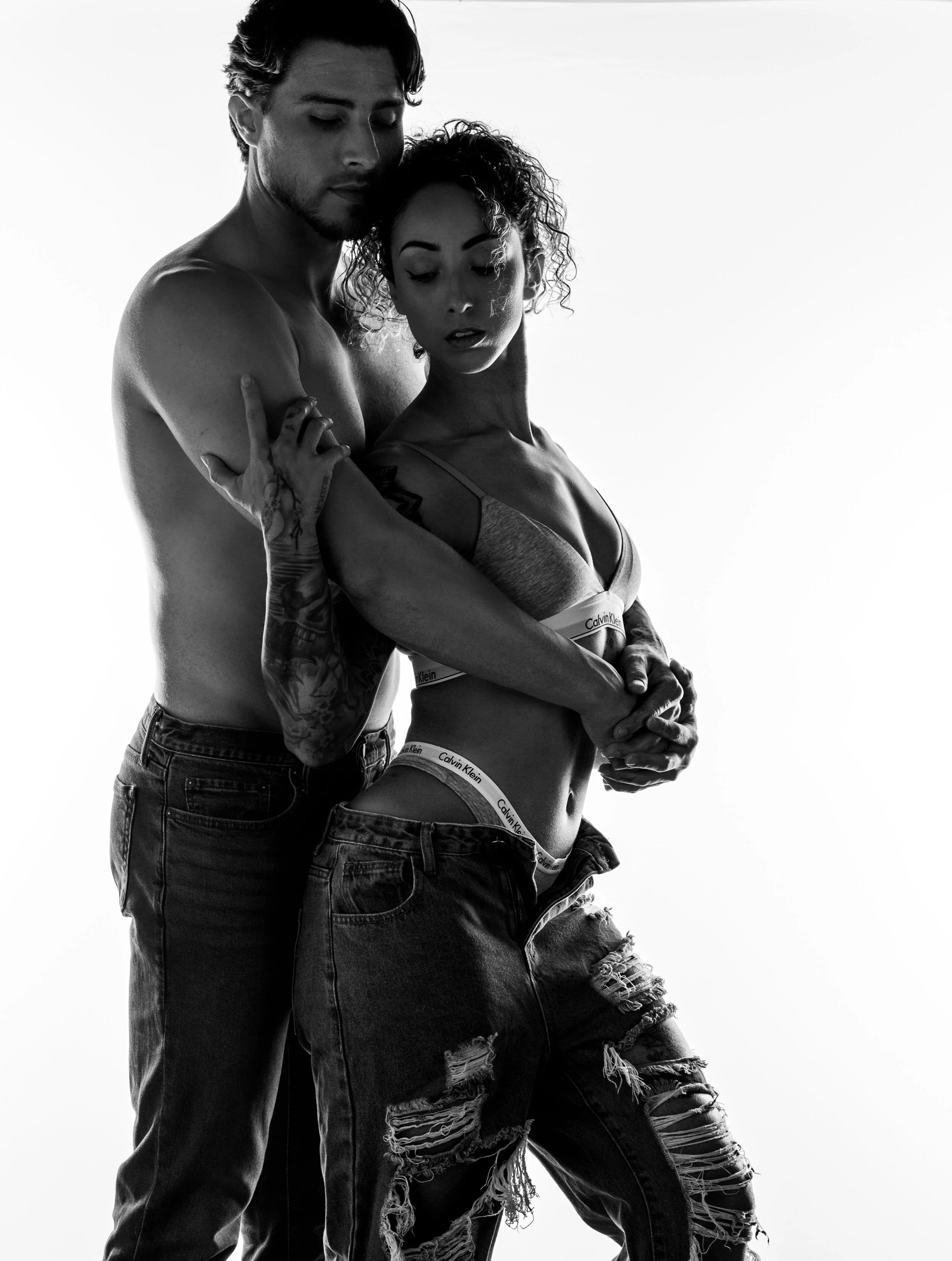 Black and white Engagement portrait of a shirtless man and woman in jeans embracing closely against a white background, with dramatic side lighting emphasizing their figures.  Couples Boudoir.