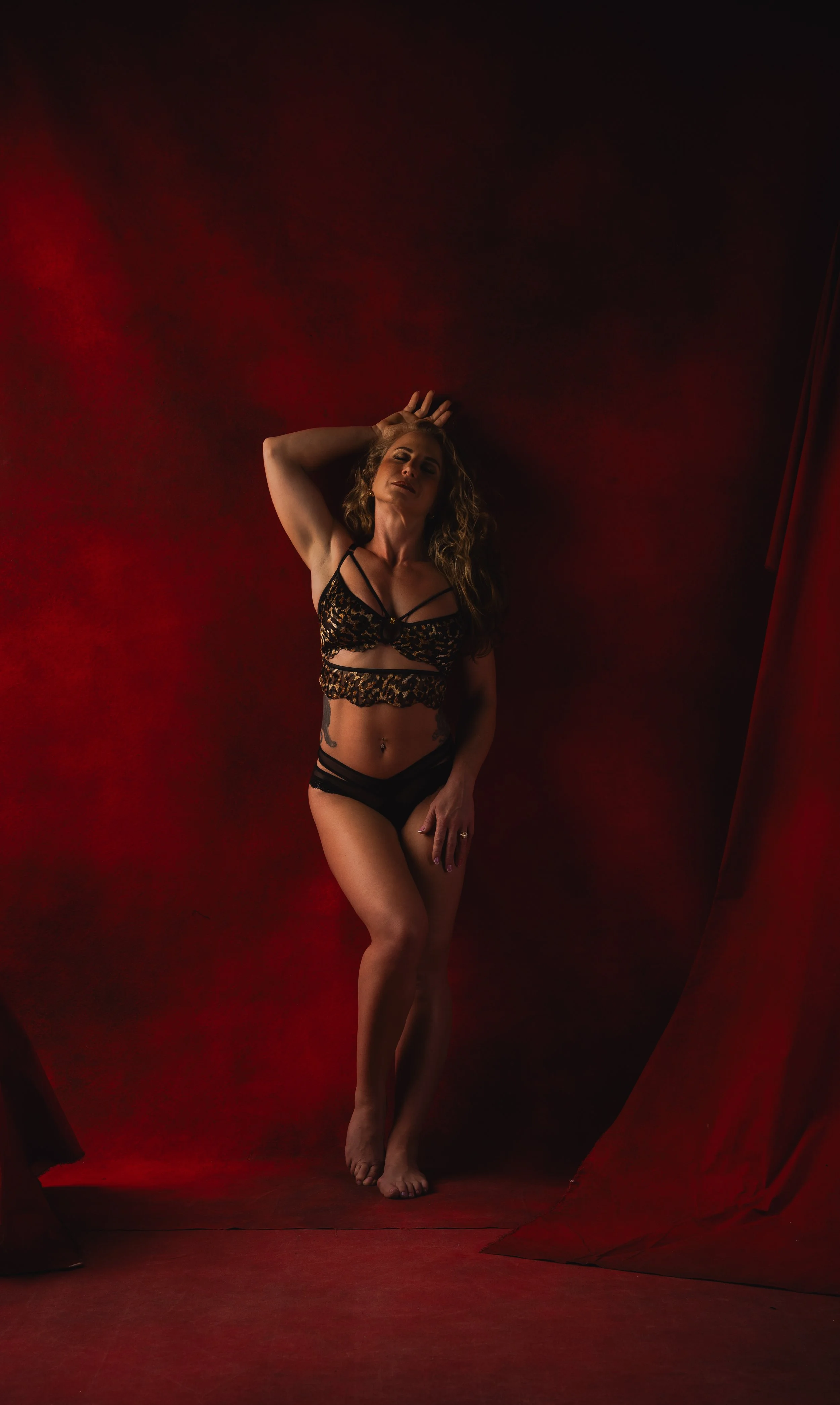 A woman with curly hair in lingerie standing against a red and black backdrop with soft lighting.