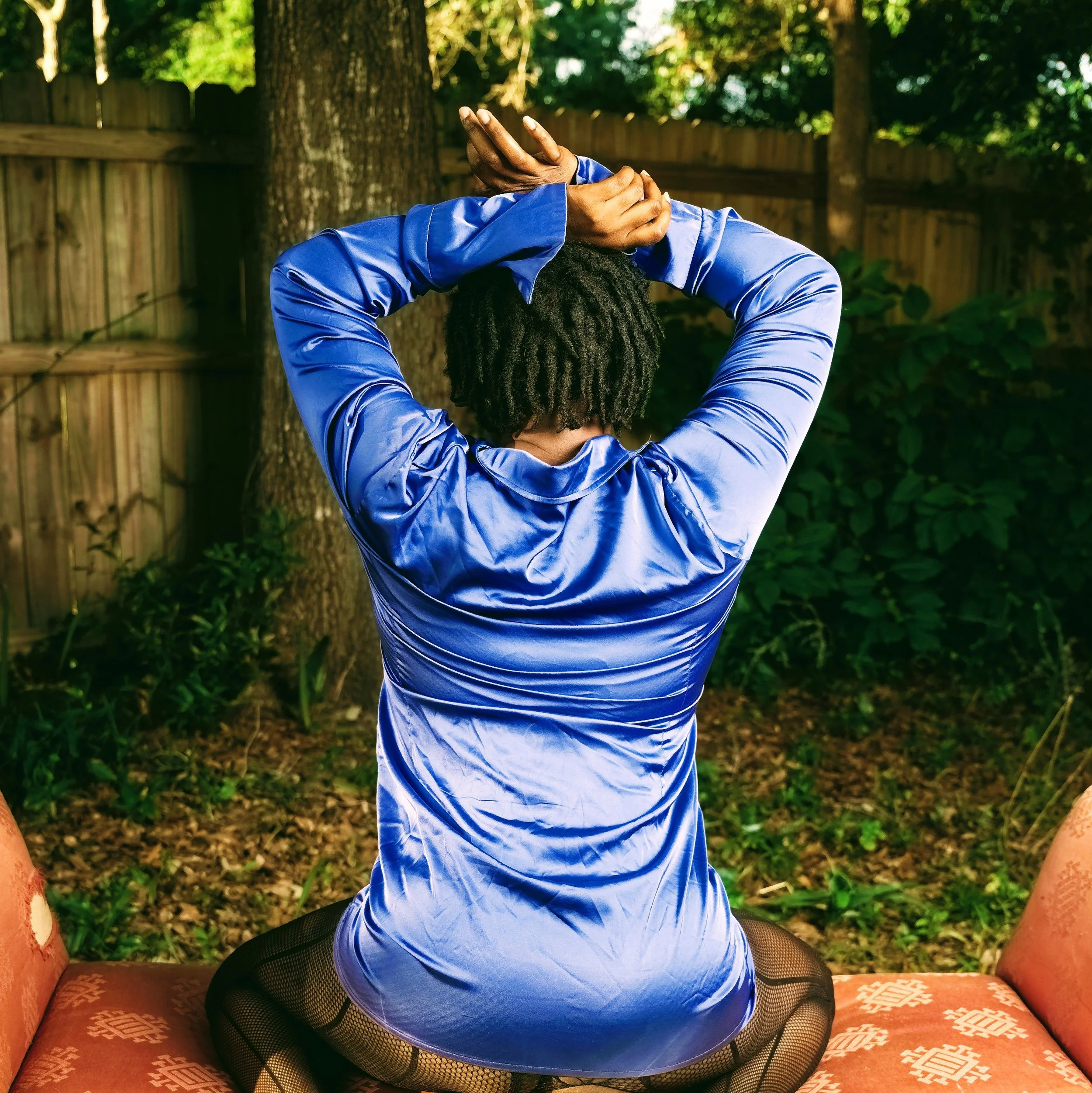A woman sitting on her spread out knees while on an orange patterned bench outdoors, wearing black stripped fishnet stockings, and a shiny blue dress with her back to the camera. Her blue dress, too short to stay over her thighs and butt as she sits,