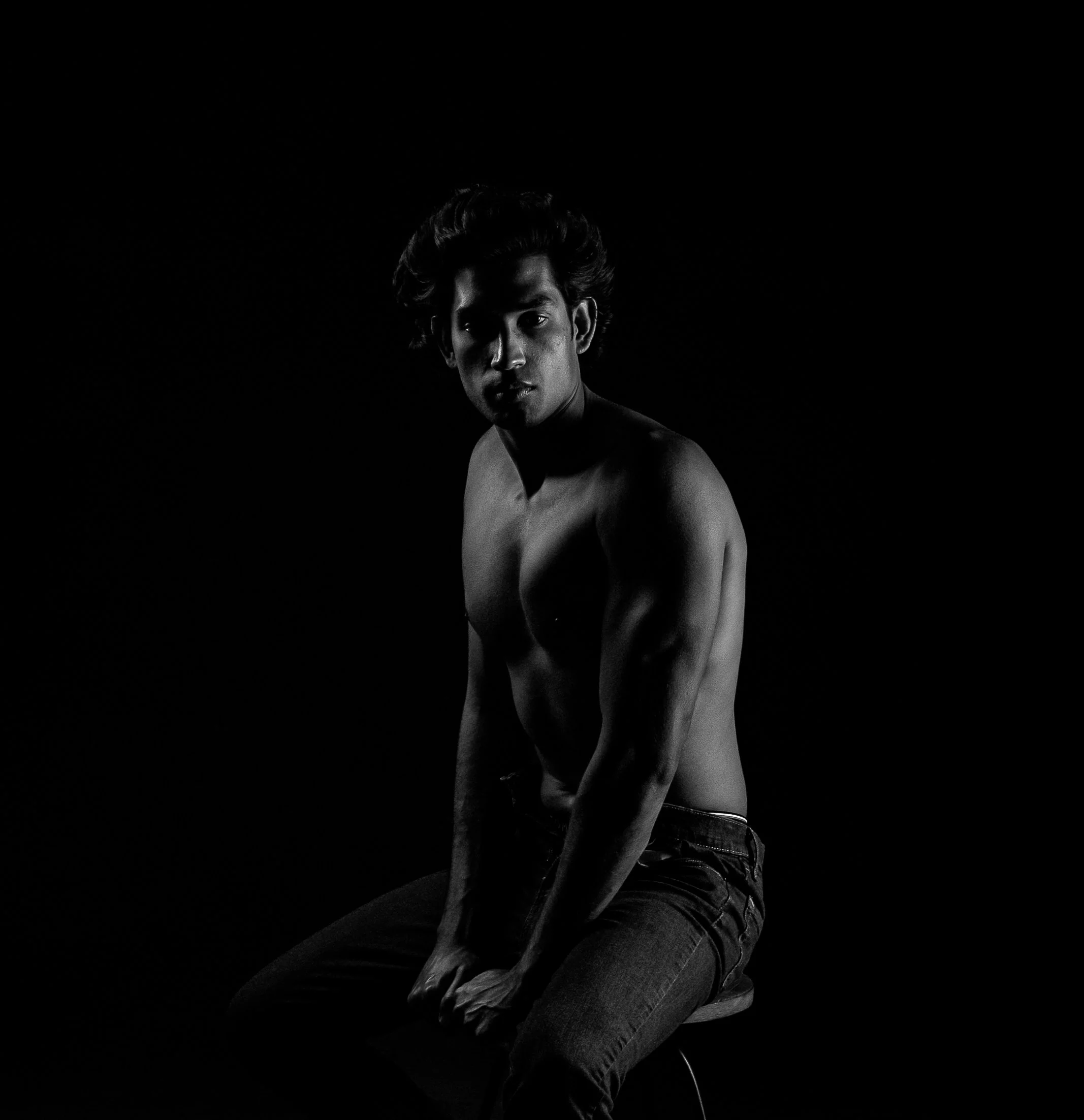 Black and white studio portrait of a shirtless young man in jeans seated and gazing directly at the camera, with dramatic low-key lighting against a pure black background.  Boudoir for Men.  Male Boudoir. Gentlemen Boudoir.  Dudeoir and masculine bou