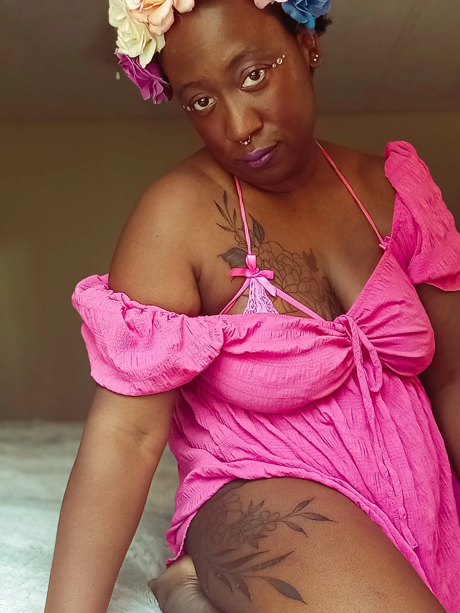 Woman with floral headband wearing pink dress, featuring a tattoo of flowers and leaves on her shoulder and thigh, posing indoors.