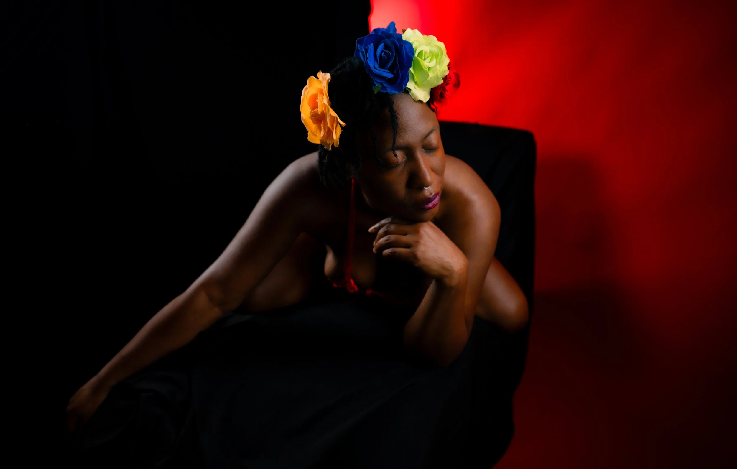 Studio portrait of a woman reclining on a dark chair with a vibrant blue, yellow, and orange floral crown, against a split black and red background with a contemplative expression.