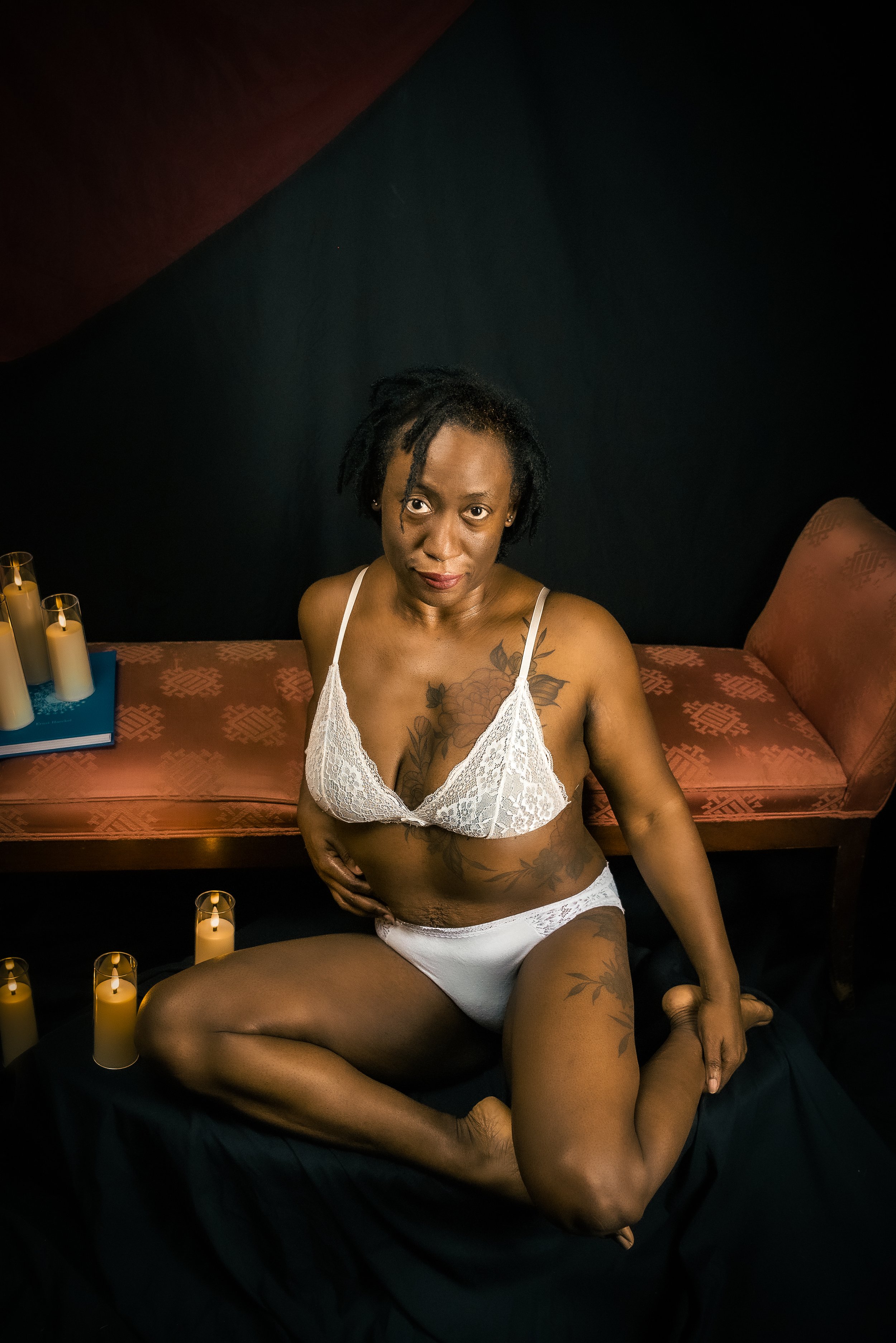 A woman with tattoos wearing white lingerie, sitting on a black cloth, surrounded by candles, in a dimly lit room with a dark background and a pinkish-red antique-style sofa.