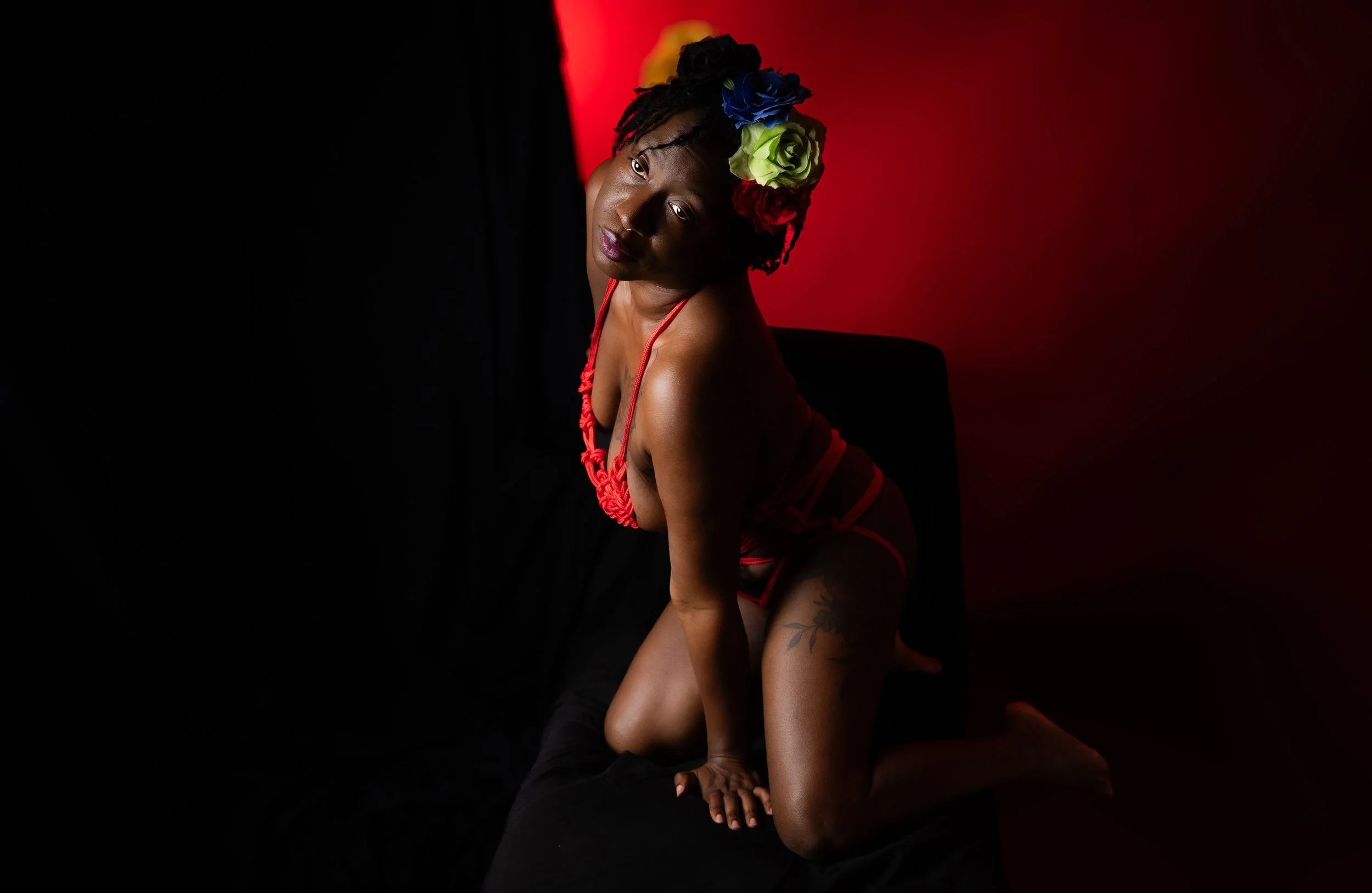 Dramatic boudoir portrait of a woman tied in red rope kneeling on a dark surface, gazing upward, with deep red lighting and floral hair accessory.  Shibari, ropework.   