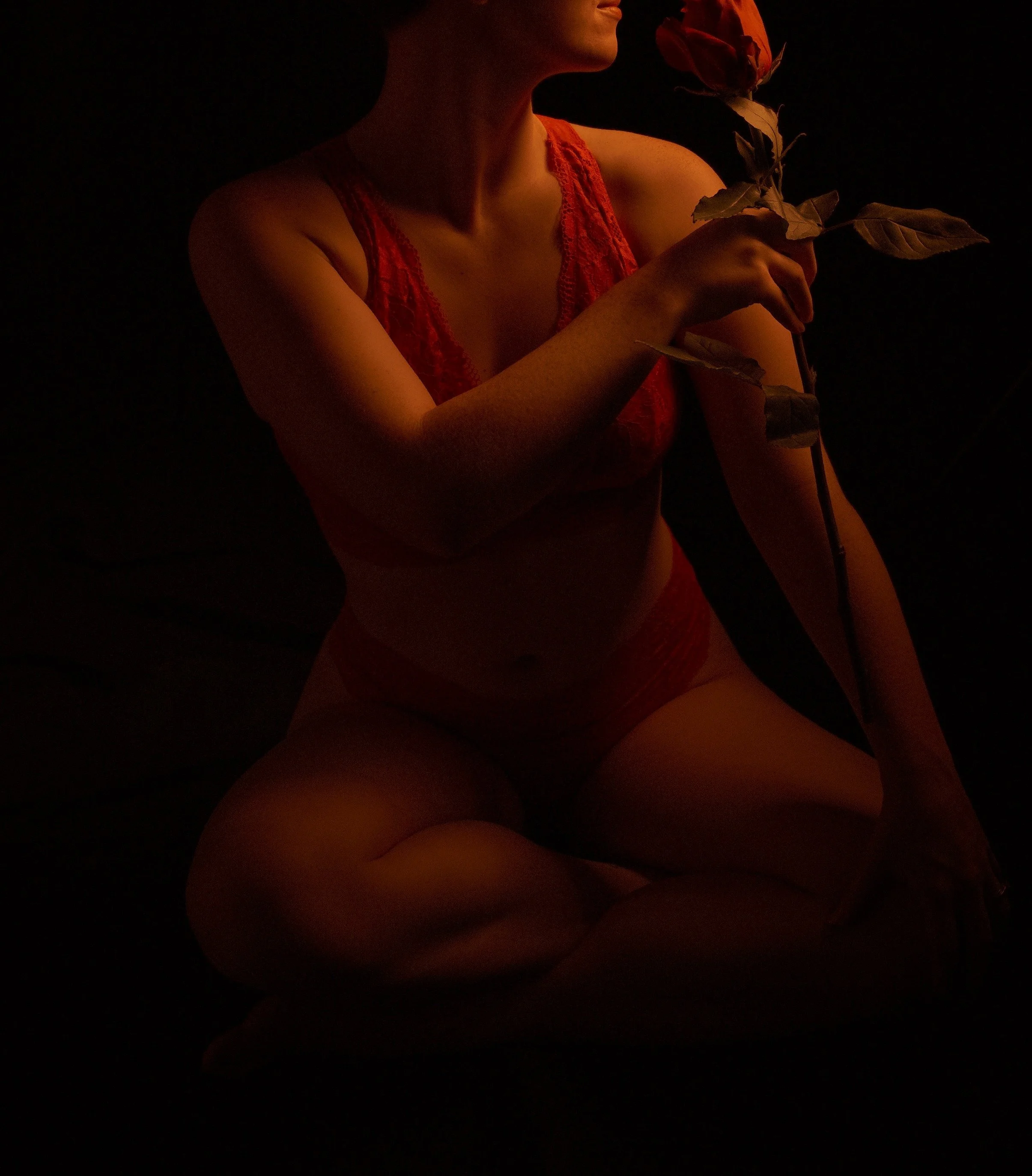A person wearing red lace lingerie sitting on the ground, holding a rose, with a dark background.