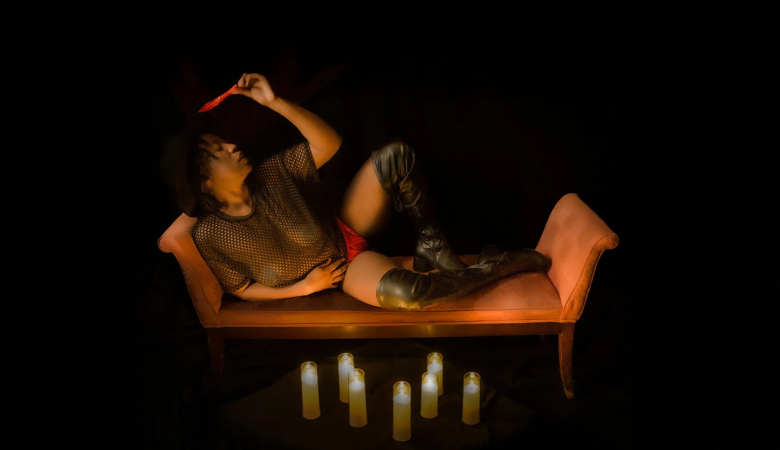 Person reclining on a vintage sofa surrounded by candles, holding a knife, with dark background.