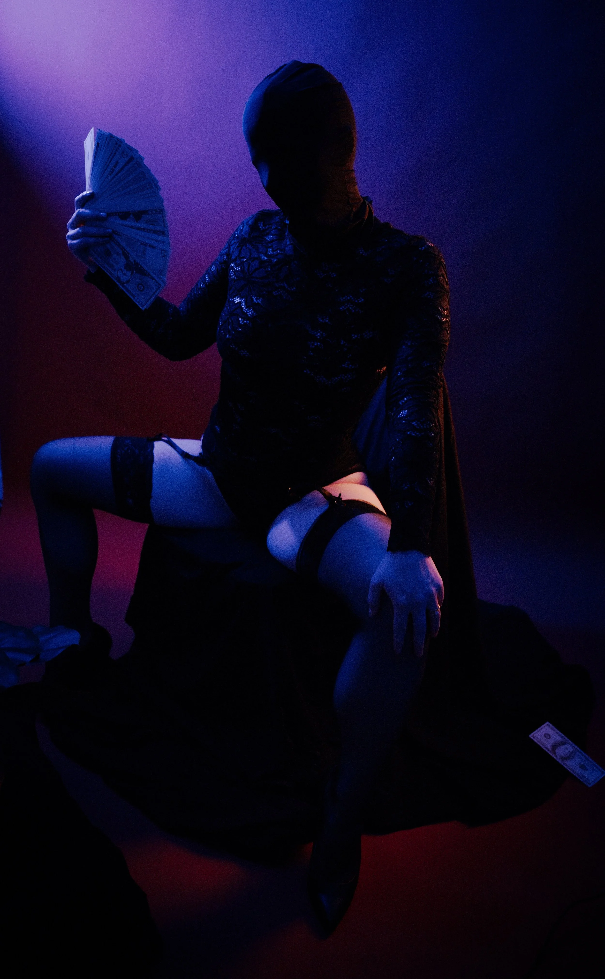A person wearing black lace clothing, stockings, and high heels, with their face obscured by a hood or mask, sitting on a surface with one knee raised, holding a fan of dollar bills, in a dark, moody setting with colored lighting.
