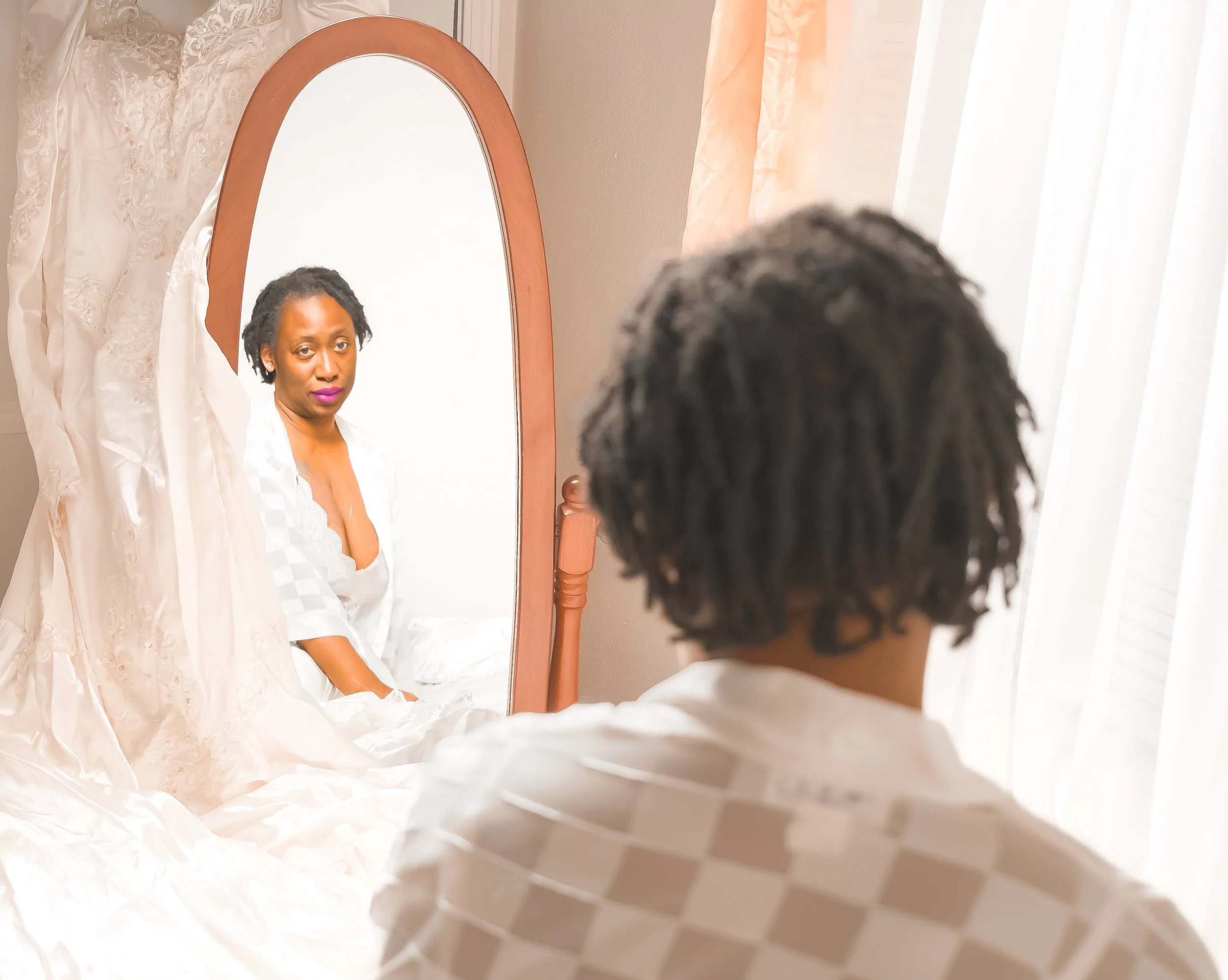 A woman with dark skin and short dreadlocks looking at herself in a mirror, wearing a white satin robe, in a bedroom with cream-colored walls and curtains.