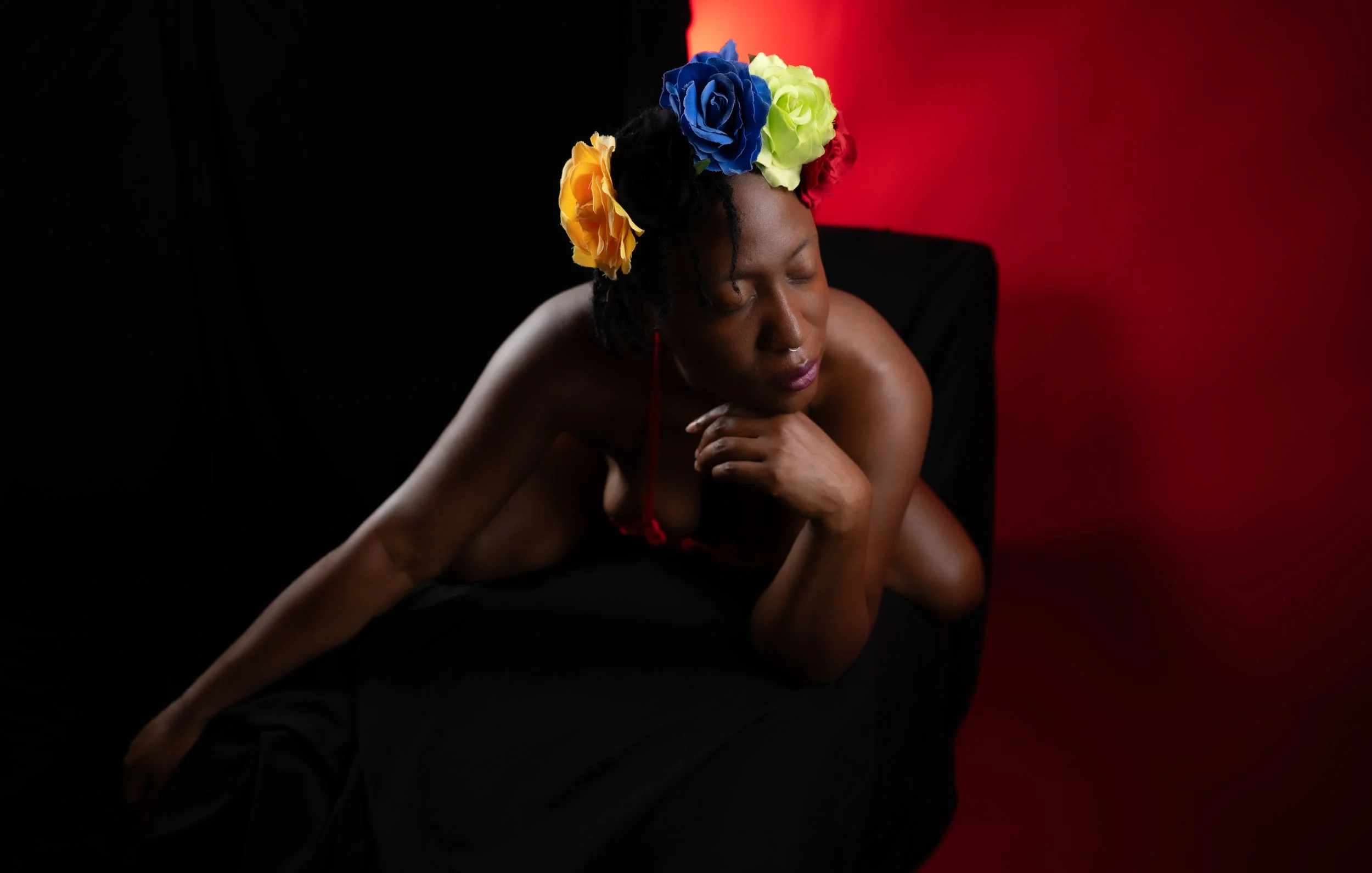 Studio portrait of a woman reclining on a dark chair with a vibrant blue, yellow, and orange floral crown, against a split black and red background with a contemplative expression.