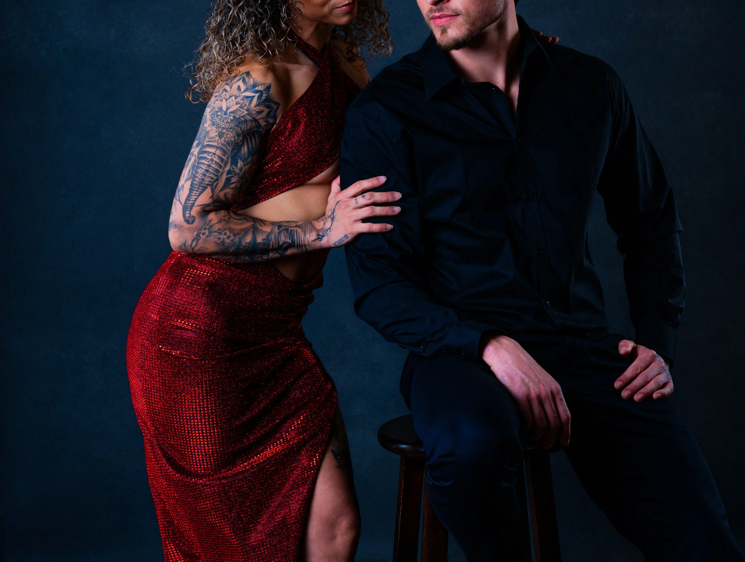 Engagement photography.  A woman with curly hair, tattoos on her arm, wearing a sparkly red dress, standing close to a man in a black shirt, against a dark background.