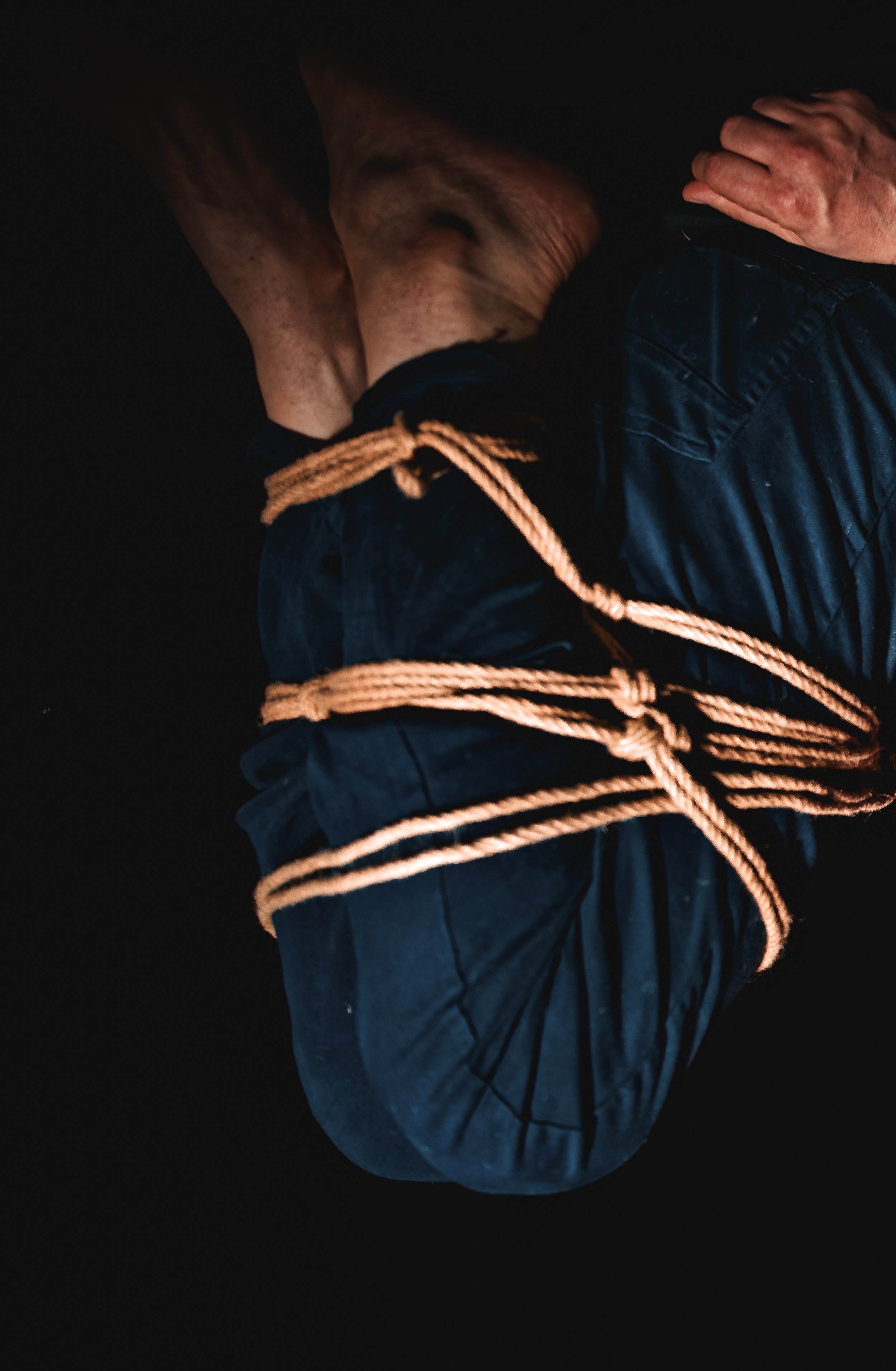 A person wrapped tightly with multiple beige ropes, with their head bowed and hands resting on their head, on a dark background.