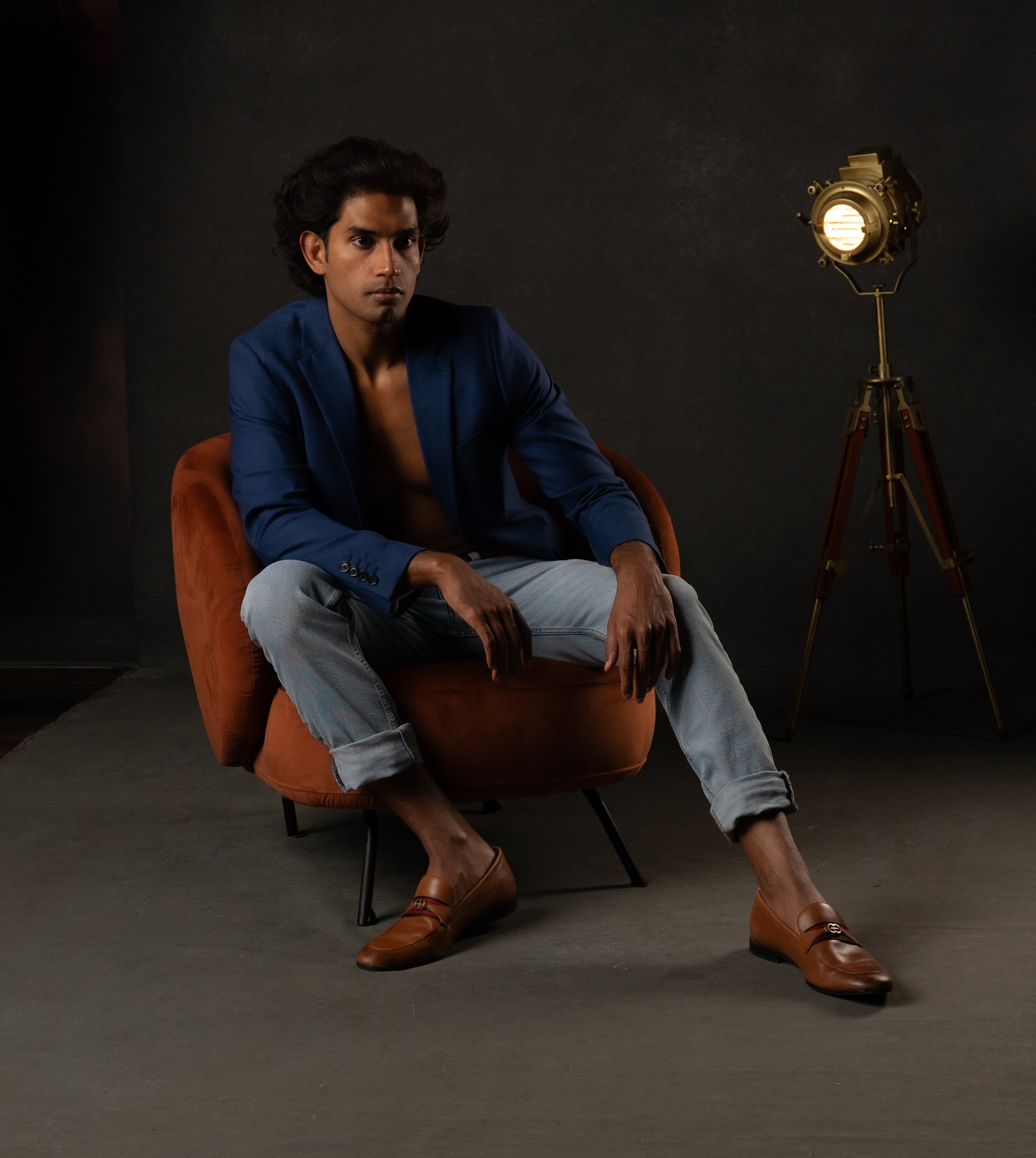 A young man with dark hair and brown skin sitting on an orange armchair, wearing a blue blazer, gray pants, brown shoes, and no shirt underneath. He is seated with his legs apart and one hand resting on his knee, looking contemplative. There is a pro