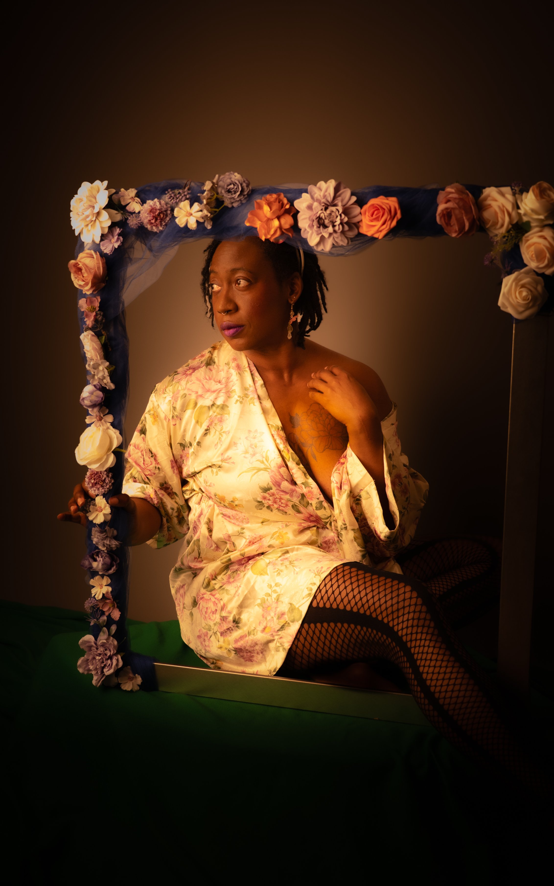 A woman with dark skin and short dreadlocks, wearing a floral silk robe, fishnet stockings, and fishnet shoes, sitting on a green platform in front of a mirror decorated with flowers. She is looking to her left with a contemplative expression, her face illuminated with soft lighting.
