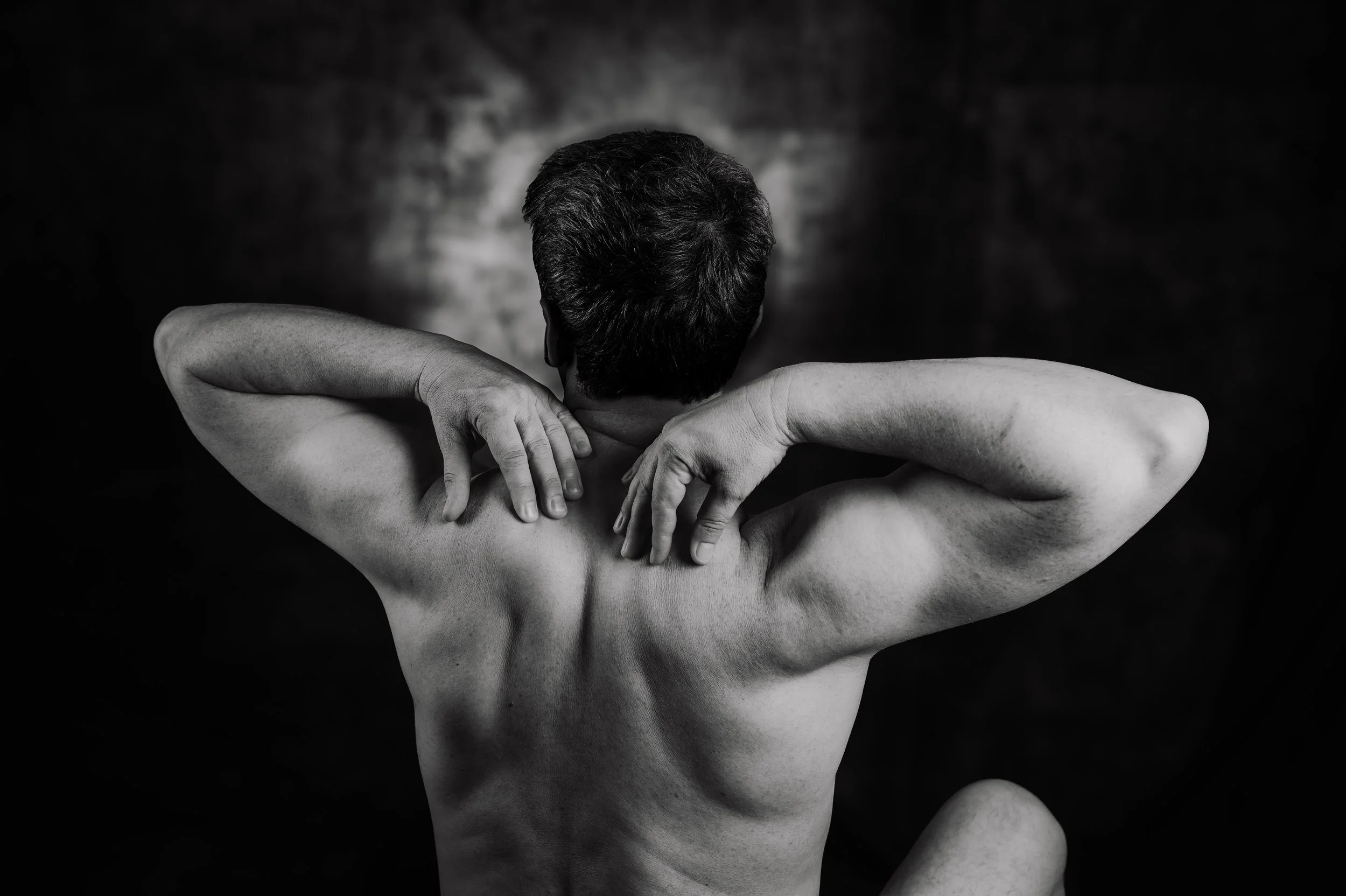 Black and white photo of a shirtless man with their back to the camera, arms crossed and hands touching their shoulders, against a dark background.  Boudoir for Men.  Male Boudoir. Gentlemen Boudoir.