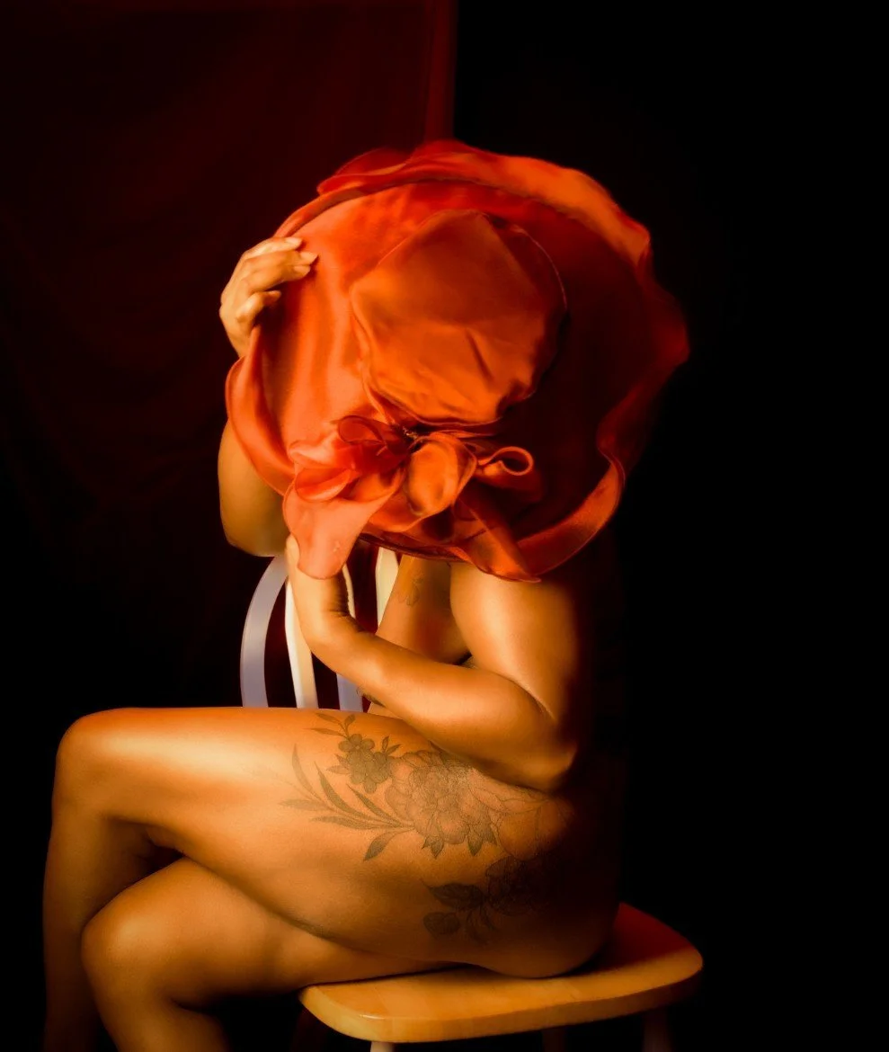 A person sitting nude on a wooden chair, with a large red satin cloth covering their head and face, holding their head with both hands, against a dark background.