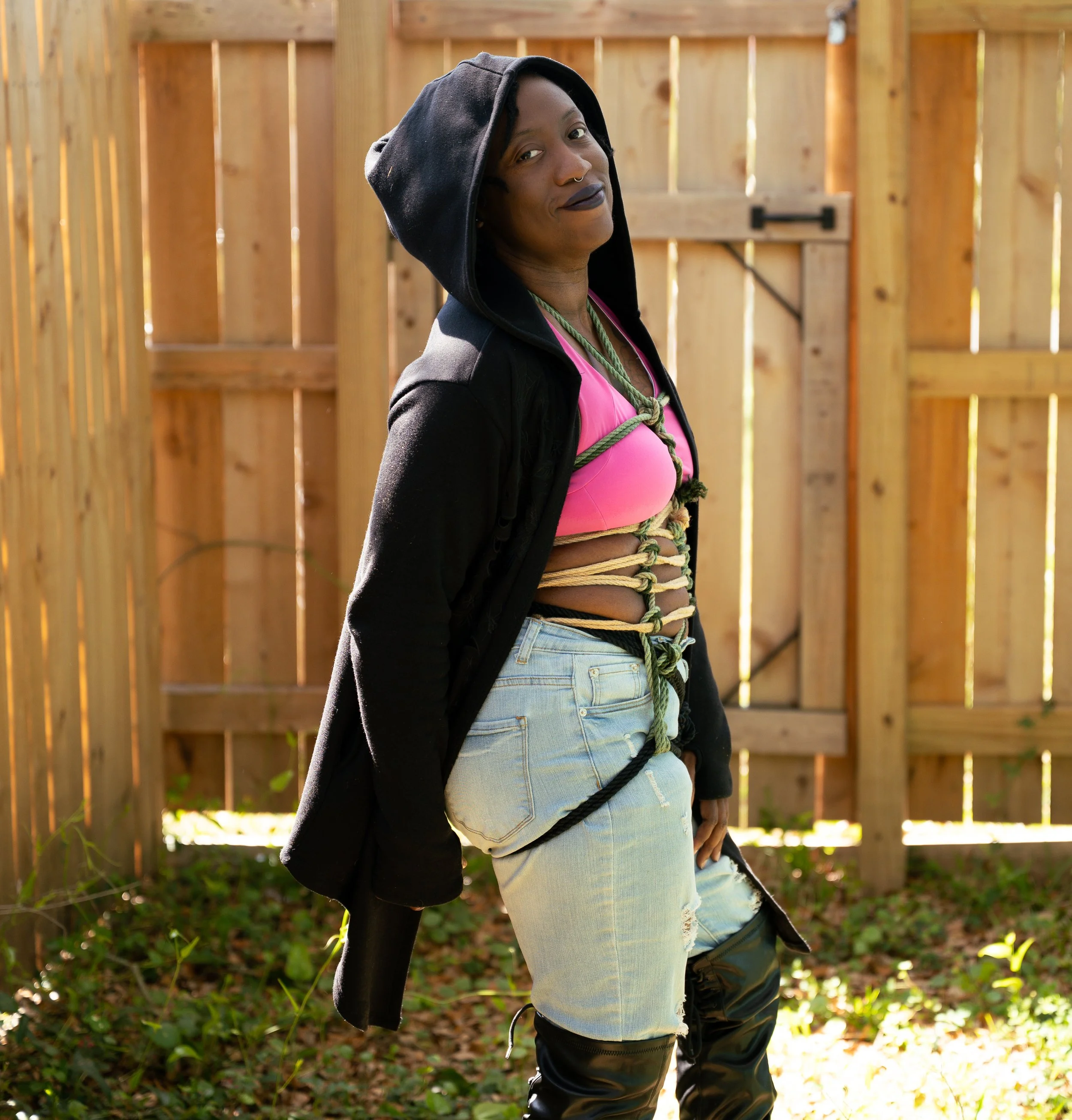 A person with dark skin and dark lipstick, wearing a black hoodie, pink top, ripped light blue jeans, and black boots, standing outdoors in front of a wooden fence, with ropes tied around their torso.