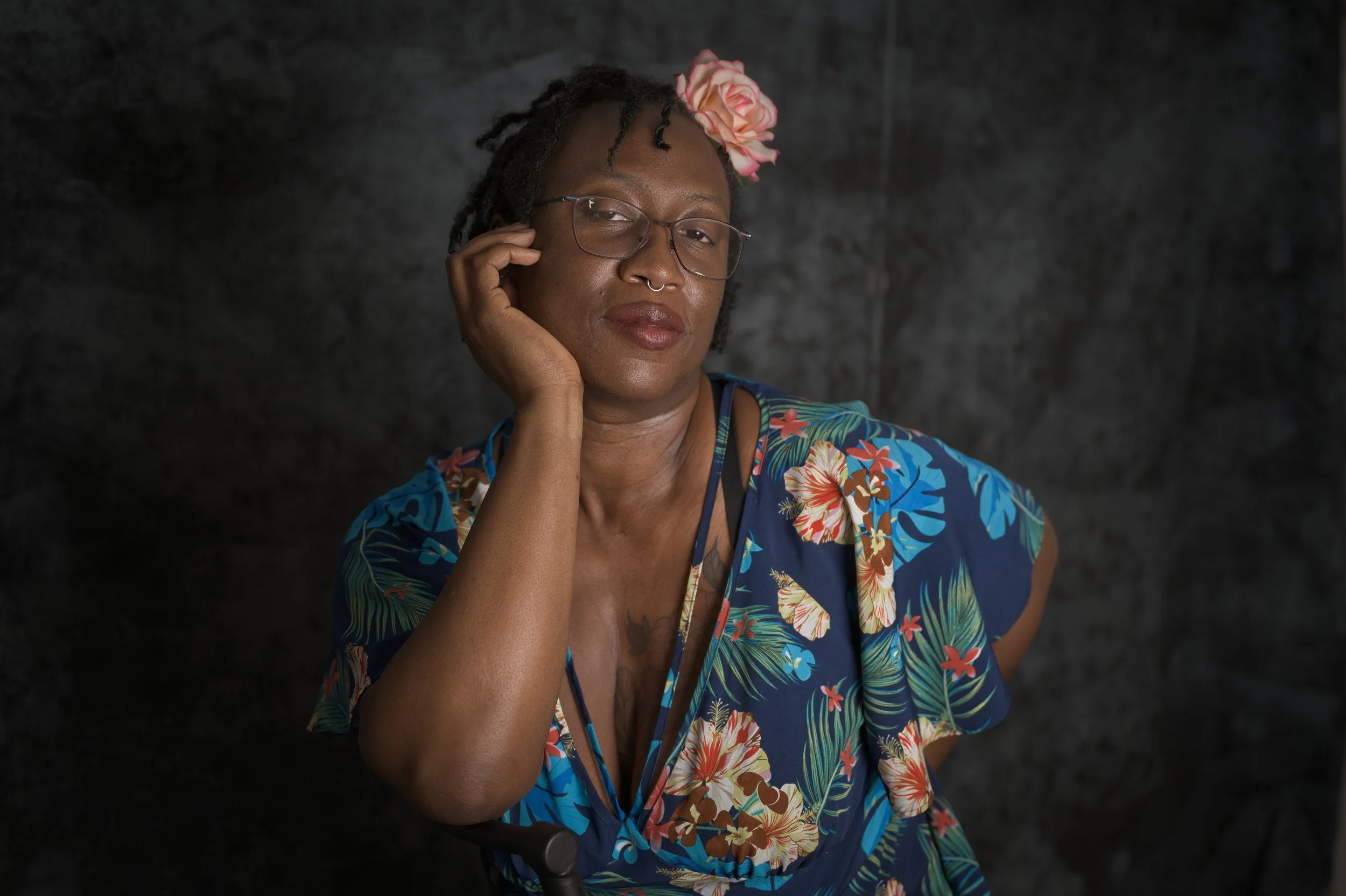 A person sitting on a chair, wearing a blue floral dress with a pink flower in their hair. Her right hand rests under the chin, while her smirky smile, and smart looking glasses create an edgy pose against a textured gray background.