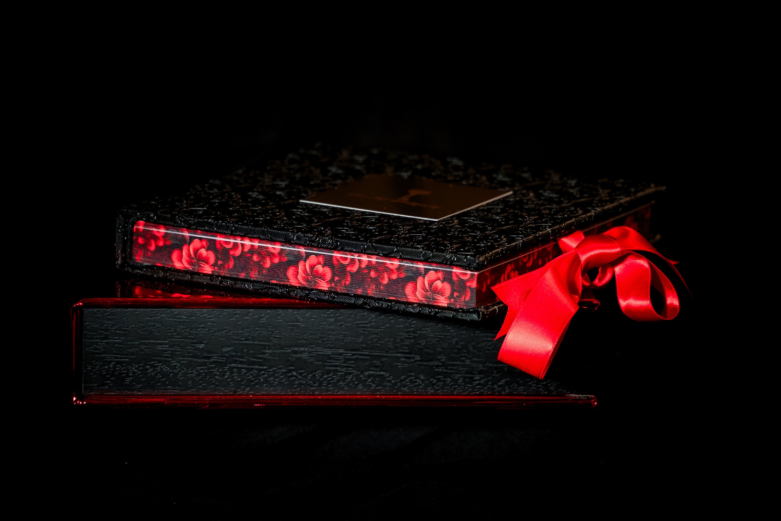 A photo box with textured surface, topped with a smaller decorated box featuring floral pattern, a shiny red ribbon tied in a bow, set against a black background.