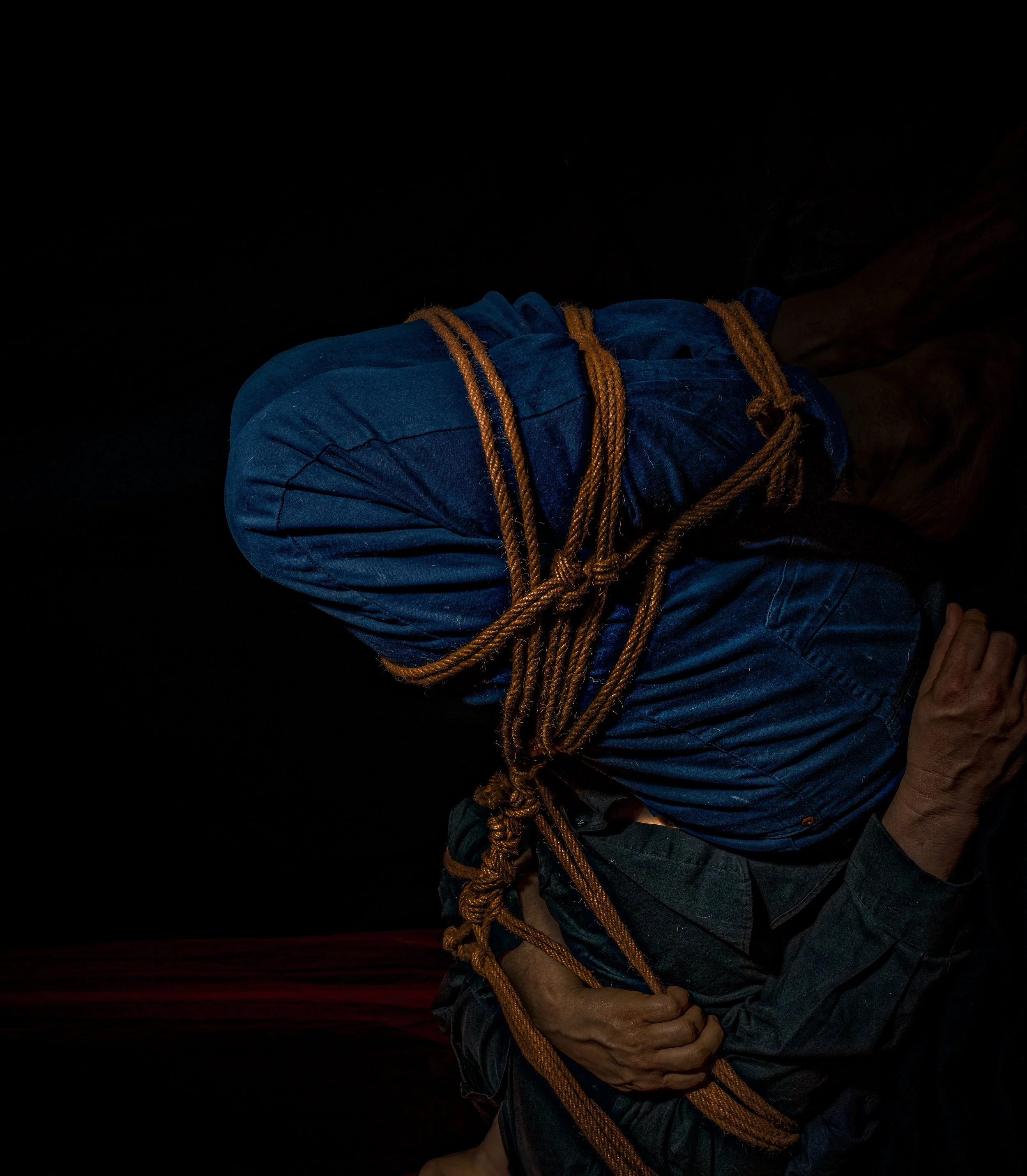 A person in a blue shirt and dark pants is tying an orange rope around themselves in a dark environment.