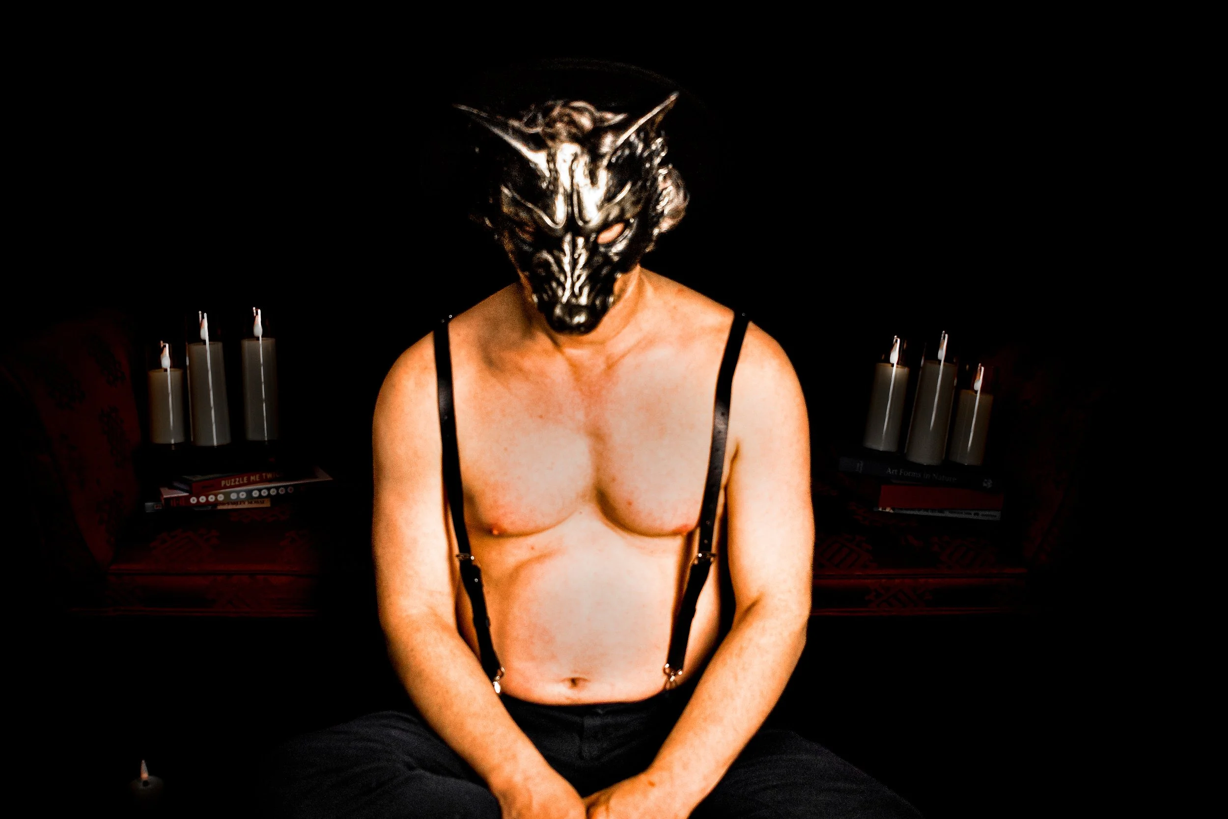 Person sitting with their shirt off, wearing a wolf mask, in a dark room with candles and books in the background.