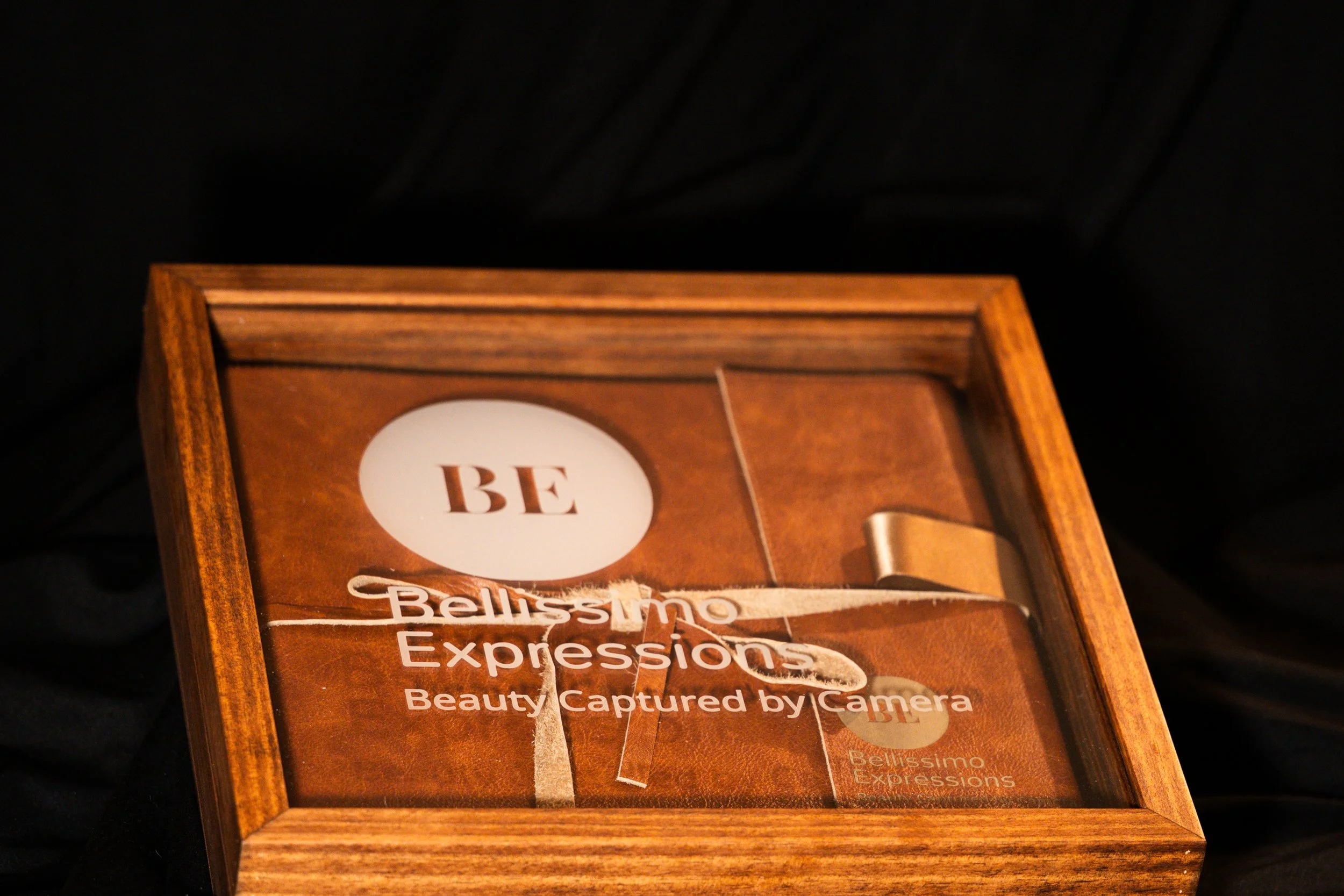 A wooden box with a glass top contains an soft textured album, and a sign inside reads 'BE Bellissimo Expressions'.
