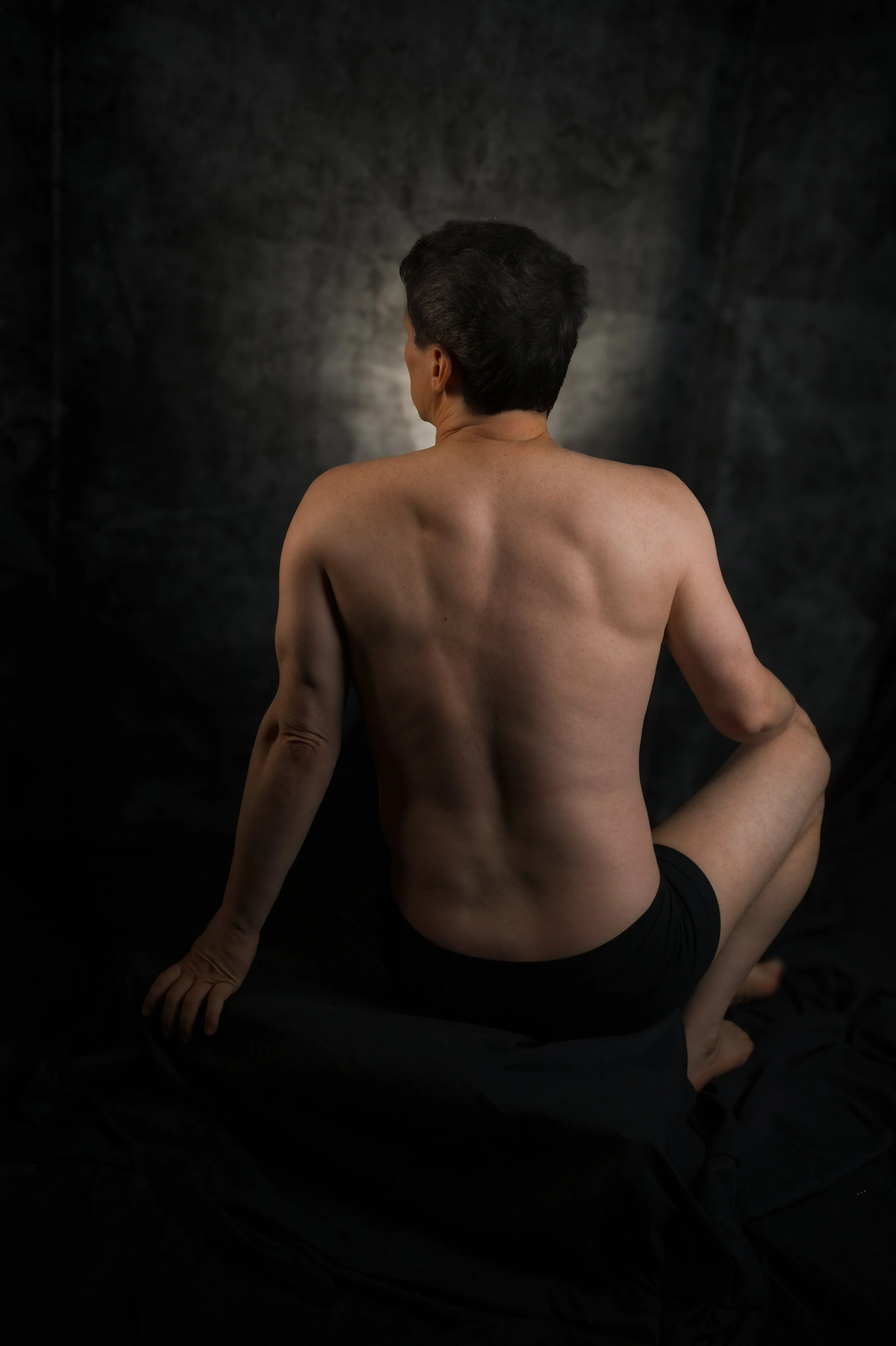 A topless man with short dark hair sitting on a black surface against a dark background. He has his back turned and is looking away from the camera.