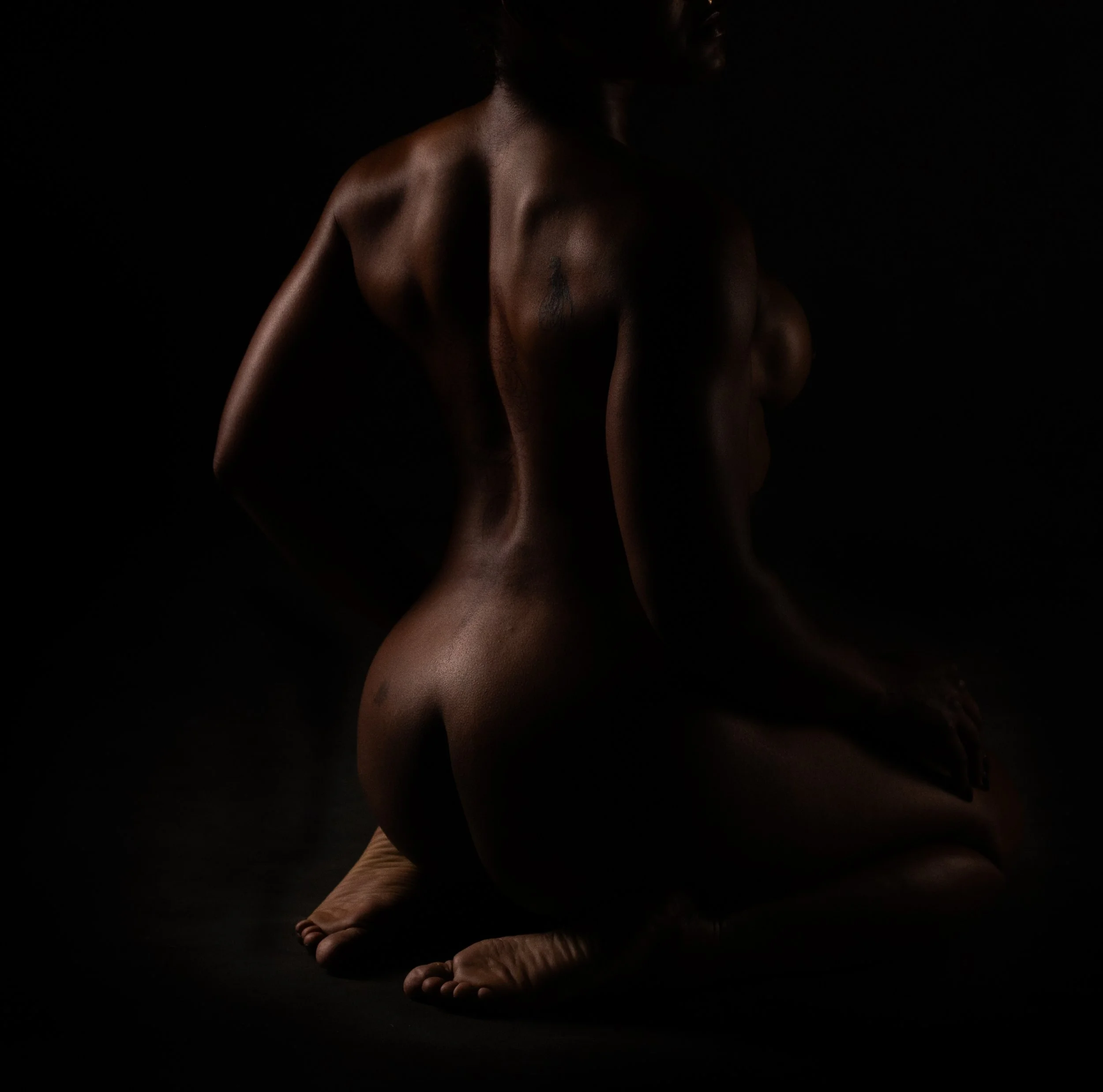 Fine art nude study of a woman seated from behind, with dramatic low-key lighting sculpting the back and form against a deep black background.