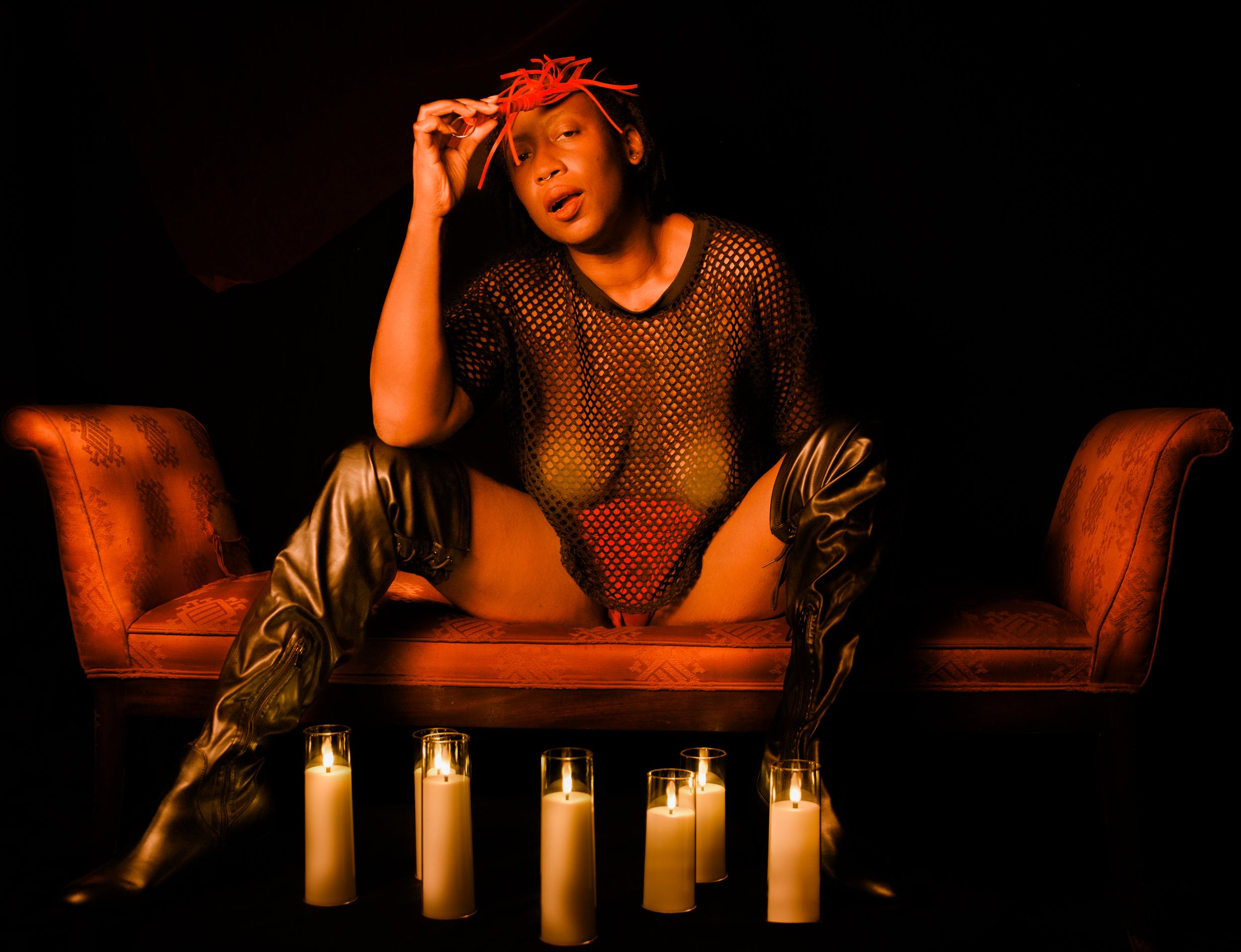 A person sitting on a vintage couch wearing a fishnet top, thigh-high boots, and a red hat, surrounded by candles in a dark setting.
