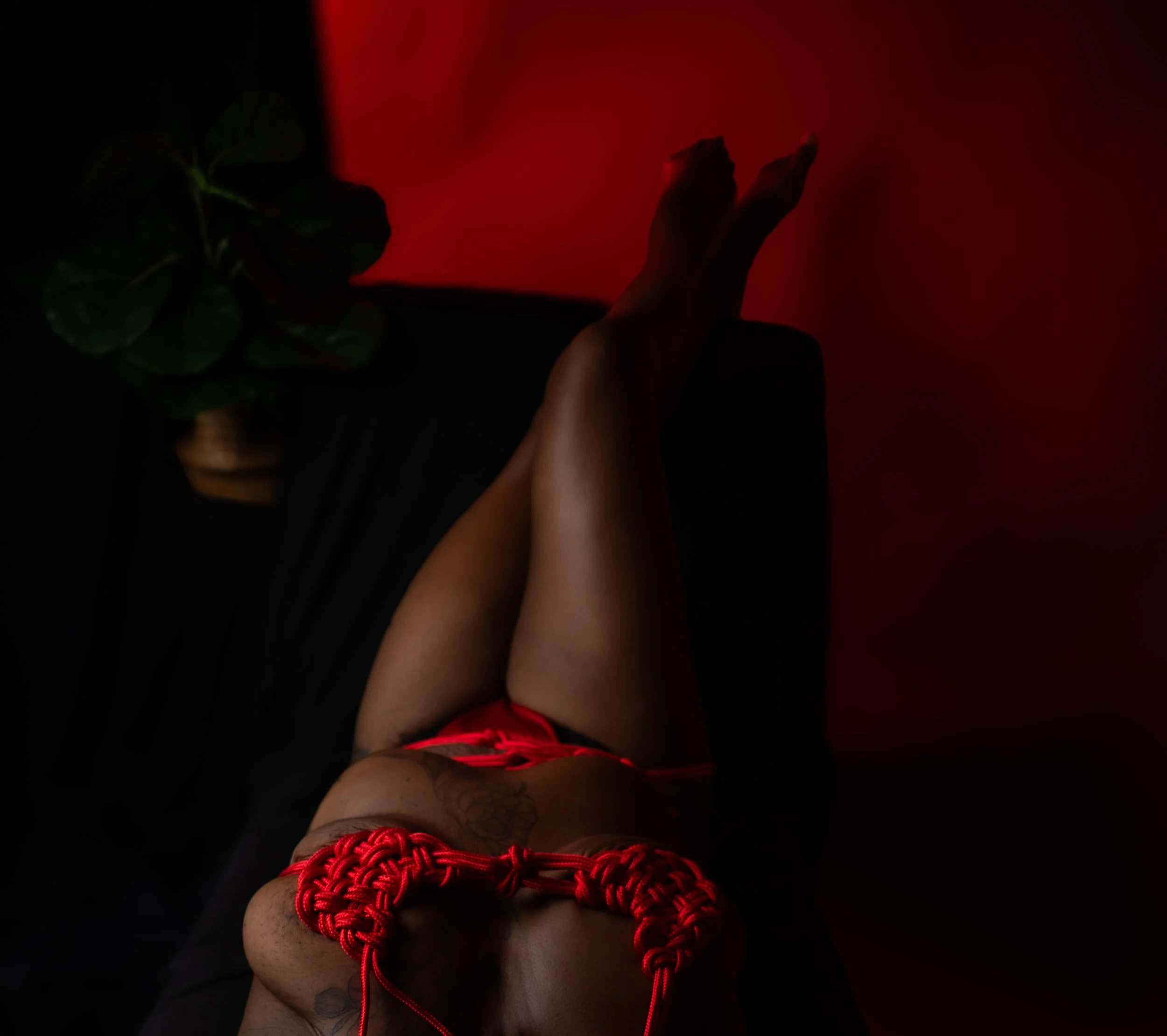 Dimly lit boudoir photo of a woman tied in red rope, bathed in red ambient lighting against a dark background, with a small circular light accent detail.  Shibari, ropework.