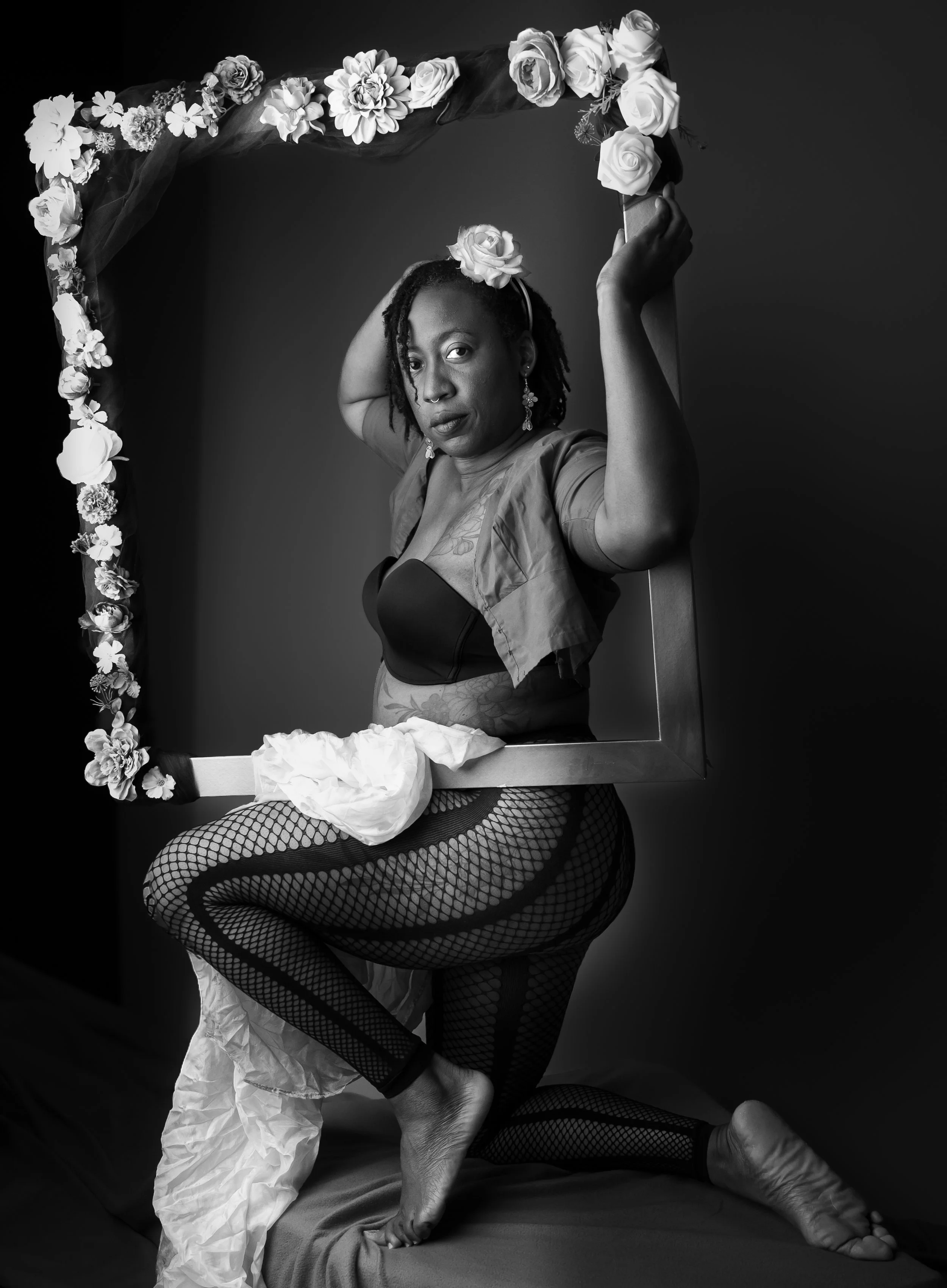 A black and white portrait of a woman with dreadlocks, sitting on a bed with fishnet stockings, holding a mirror decorated with flowers above her head. She has a confident expression and is wearing earrings and a nose ring.