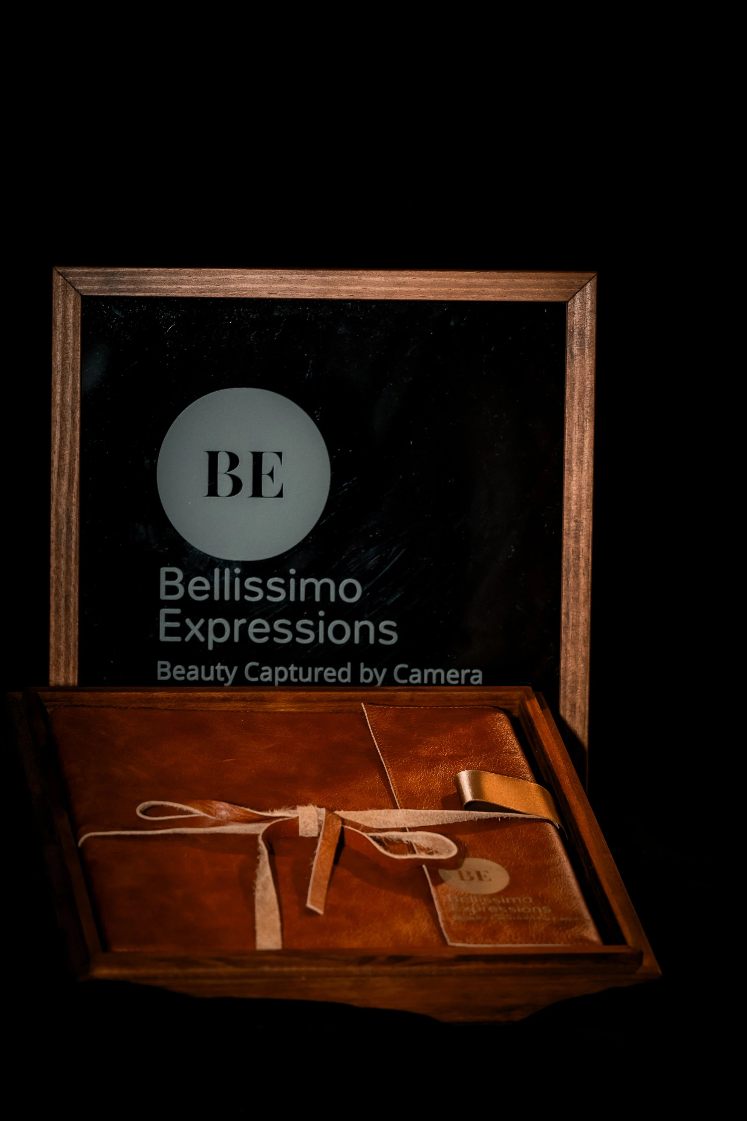 A phot box etched with the a personal engraving that says 'BE Bellissimo Expressions Beauty Captured by Camera' and a wooden box containing a brown leather item tied with a white string and a tag marked 'BE'.