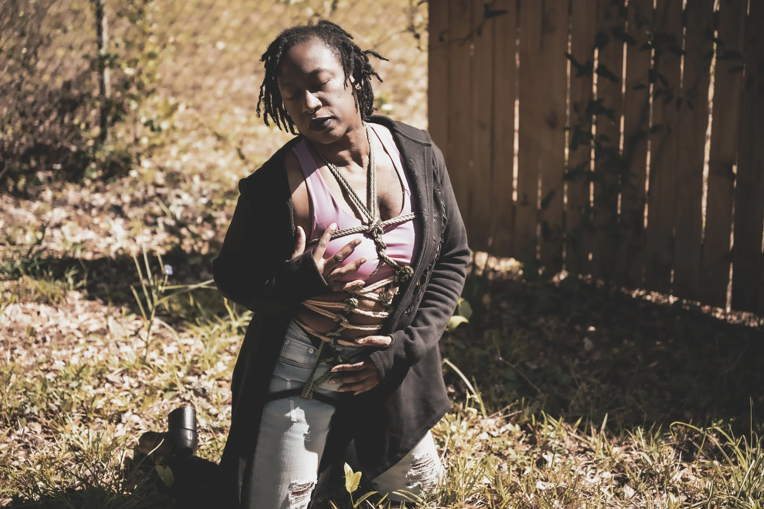 A woman with dreadlocks in a black jacket and pink top is kneeling outdoors, tied up with rope, with her hand on her chest and one on her stomach, in front of a wooden fence and surrounded by fall foliage.