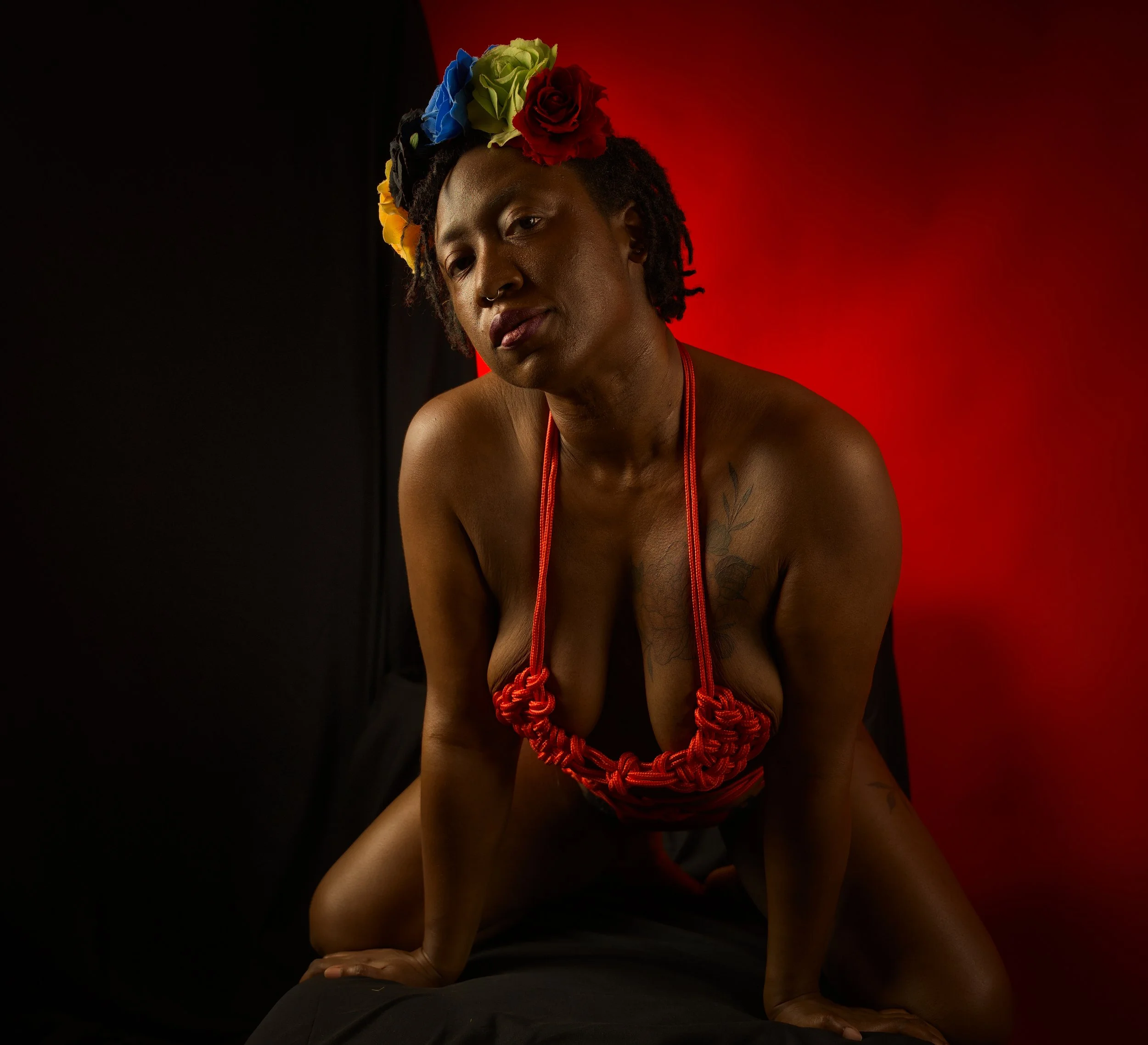 A topless woman with a floral headpiece, red necklace, and tattoos, kneeling on a black surface in front of a red and black background.