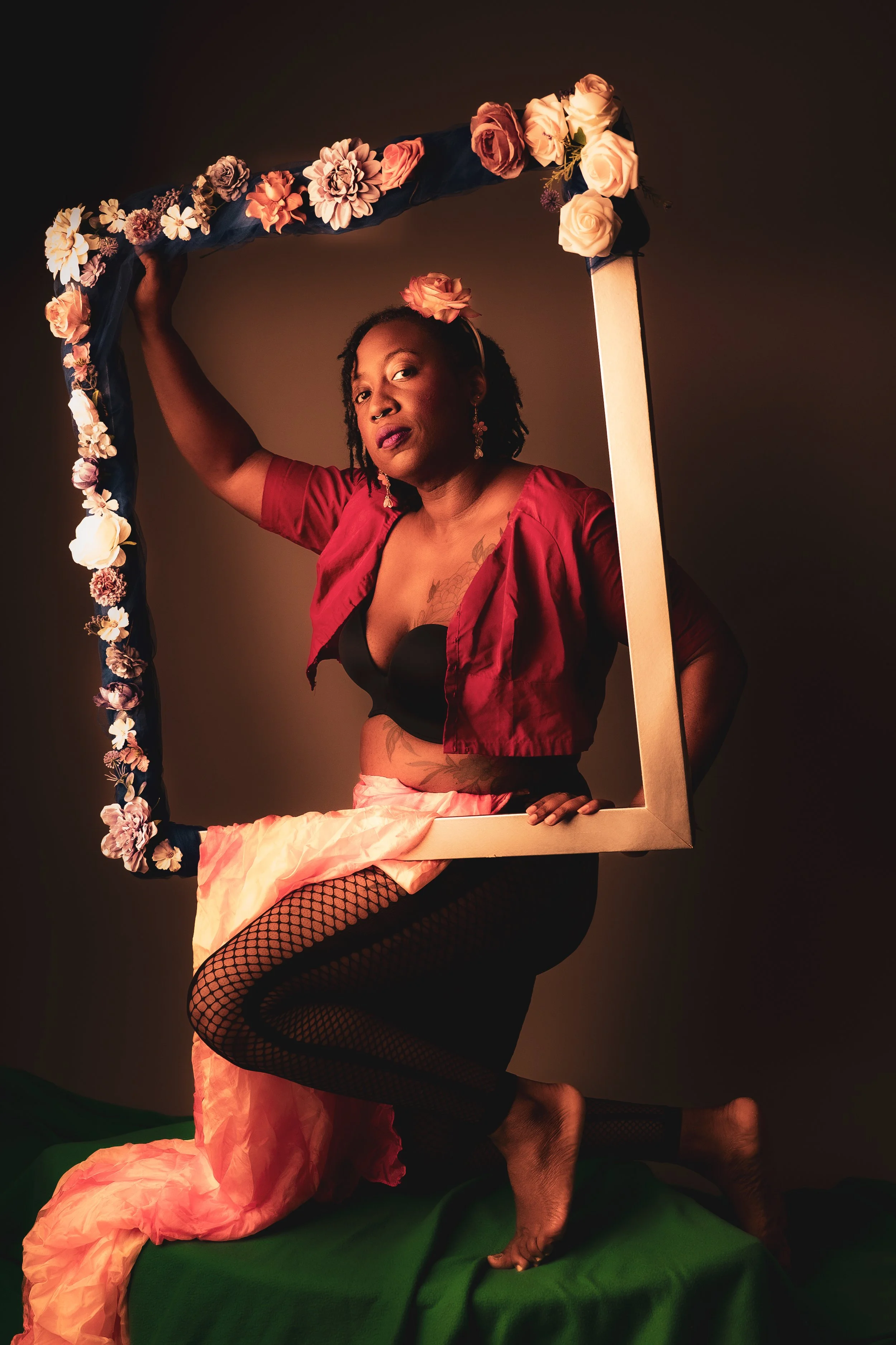 A woman with dark skin, dreadlocks, and floral earrings posing with a large floral frame and a piece of pink fabric draped over her lap. She is wearing a red crop top, fishnet stockings, and has a flower in her hair, standing against a plain dark bac