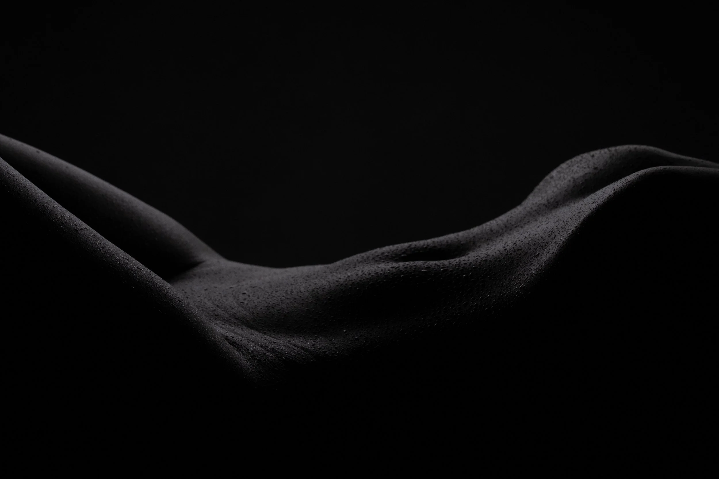 Abstract fine art bodyscape of the curved female form with dramatic low-key lighting, emphasizing contour and texture against a black background.  Bodyscape.  Fine Art Nude.