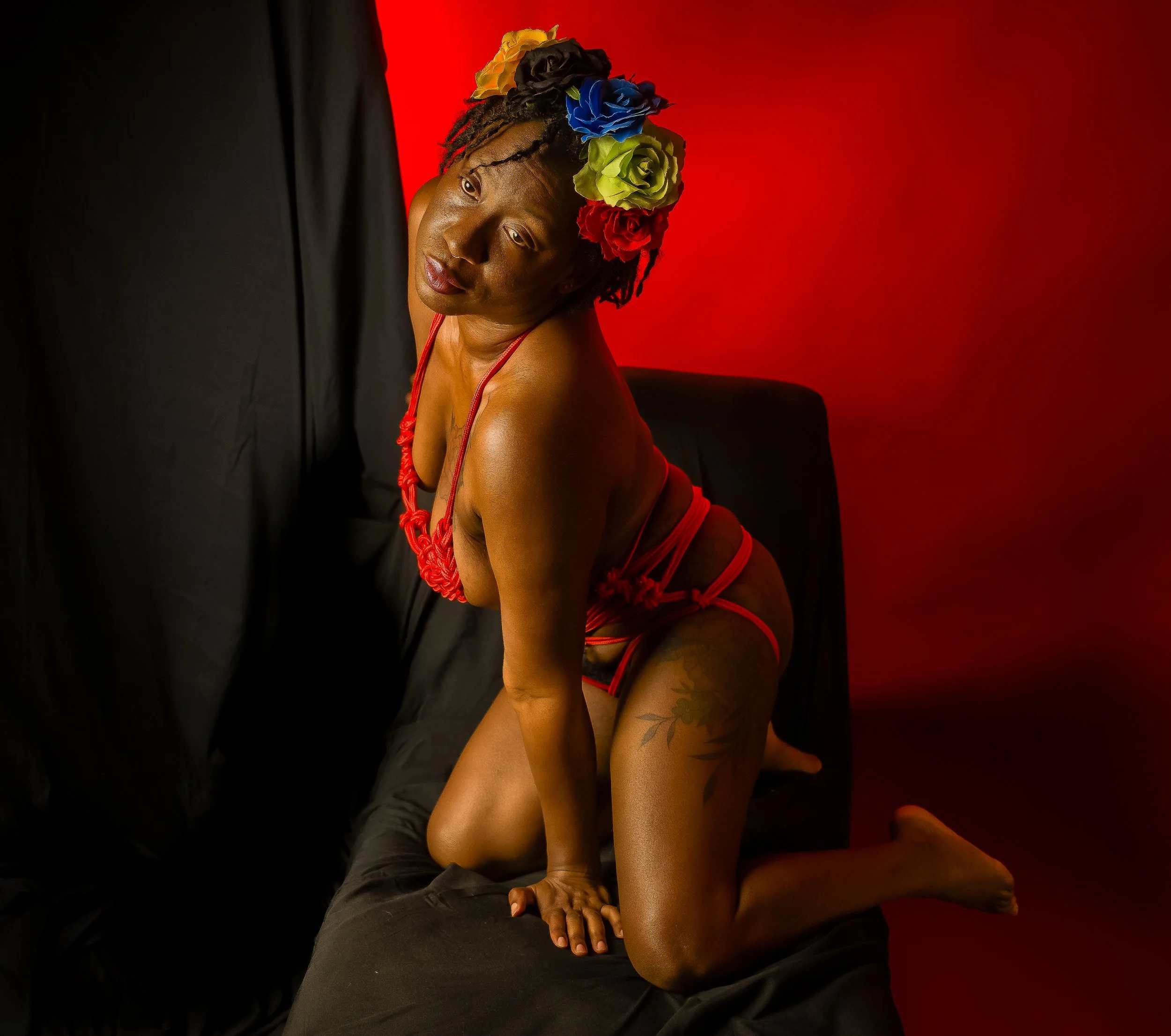 Woman wearing a colorful flower crown and red clothing, kneeling on a black couch against a red background.