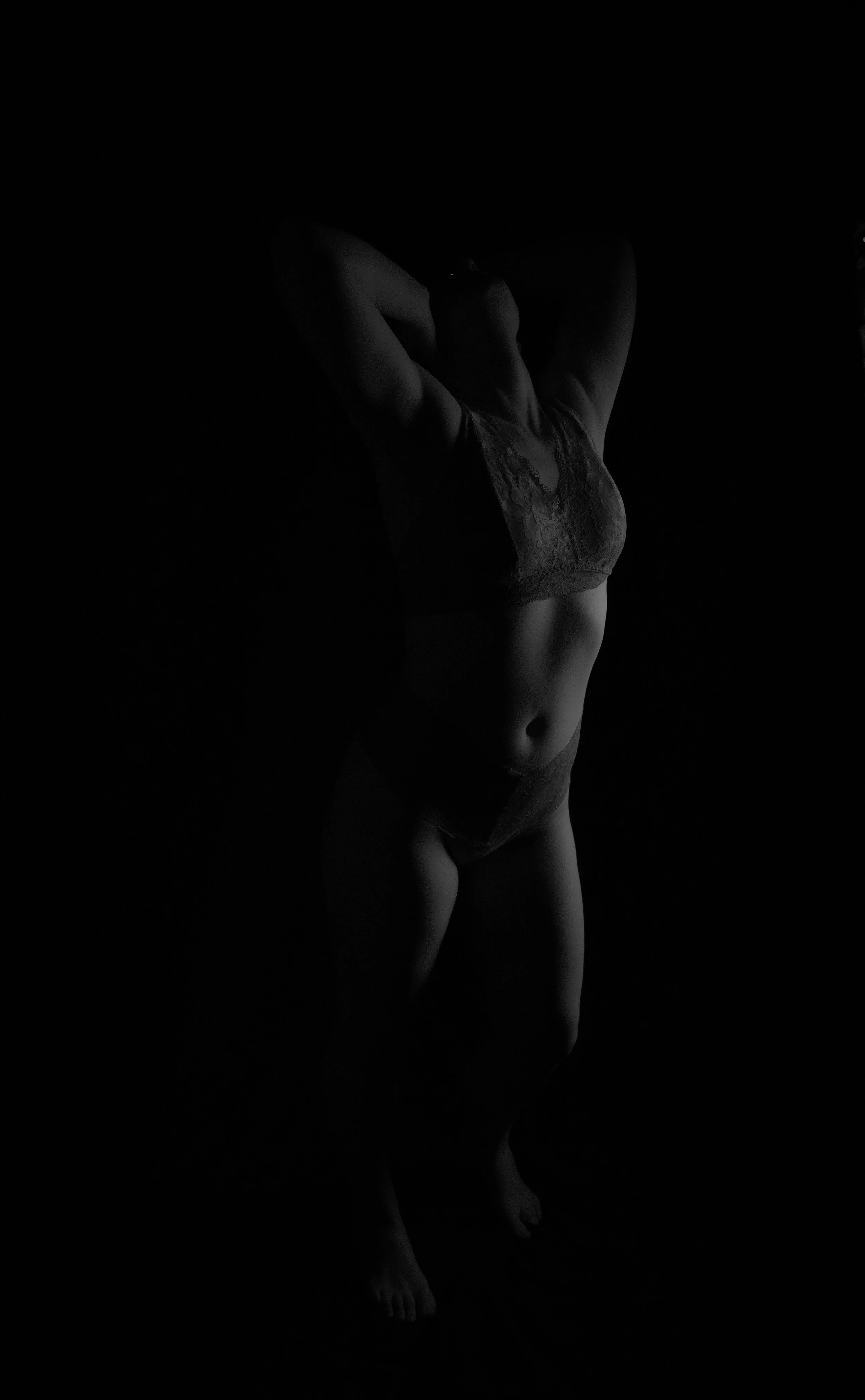 A woman is standing with her hands behind her head, wearing lingerie, in a dark setting with low lighting.