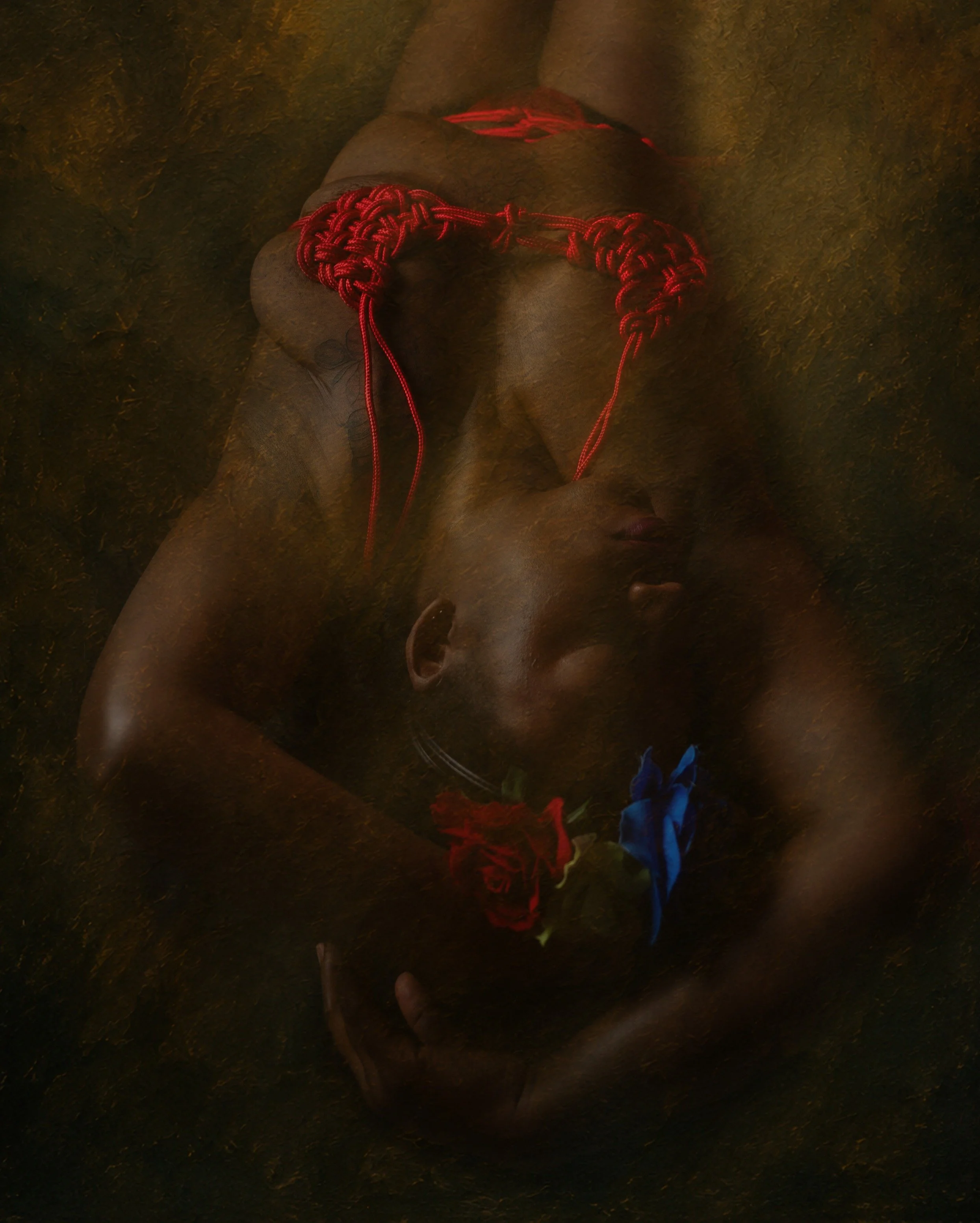A person lying on their back on a dark surface with red, blue, and yellow flowers placed on their head and red rope tied around their chest and waist. The person's eyes are closed, and they are shirtless, with a red string tied around their chest.