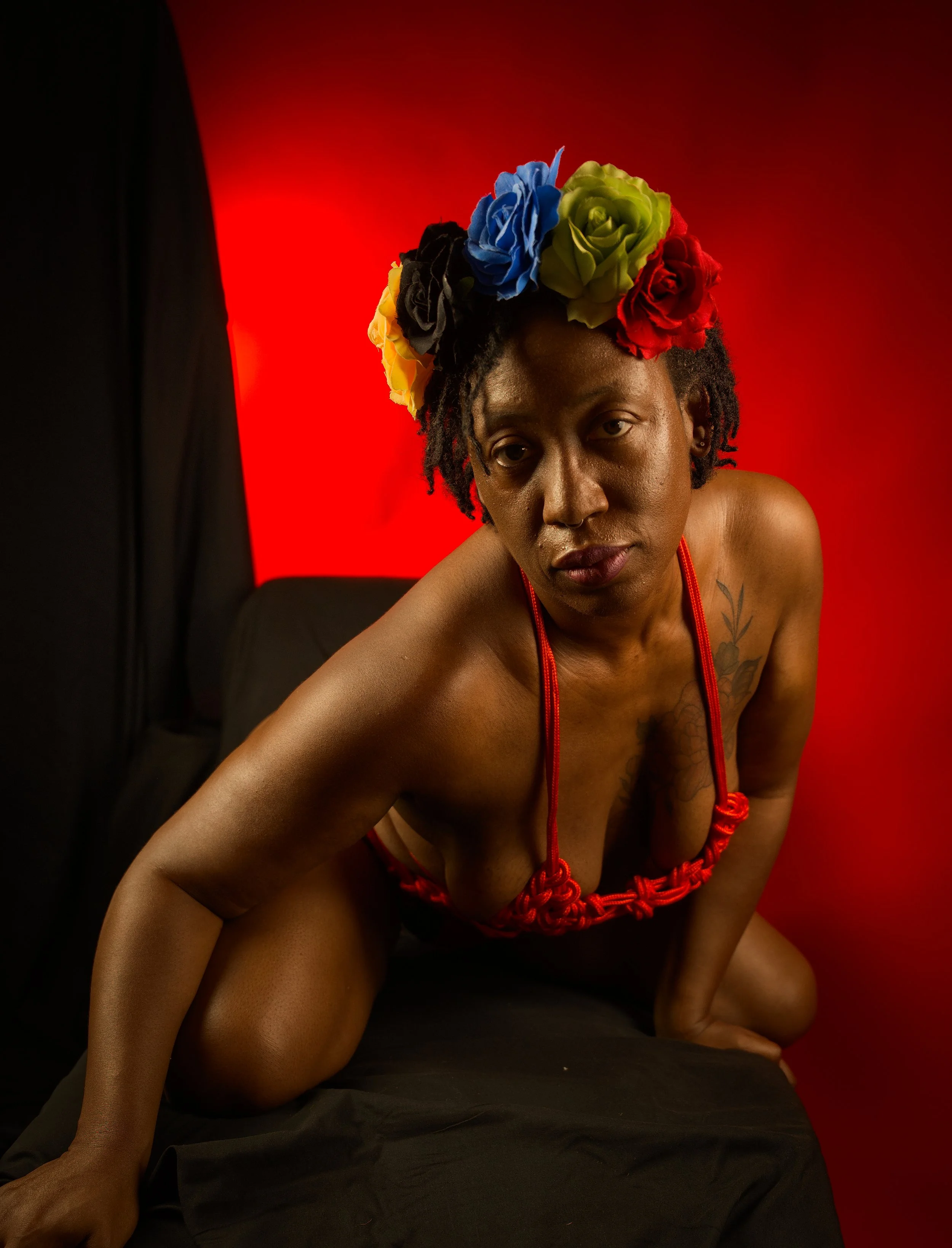 A woman with dark skin and short curly hair wearing a colorful flower crown and red necklace, posing on her hands and knees against a red background.